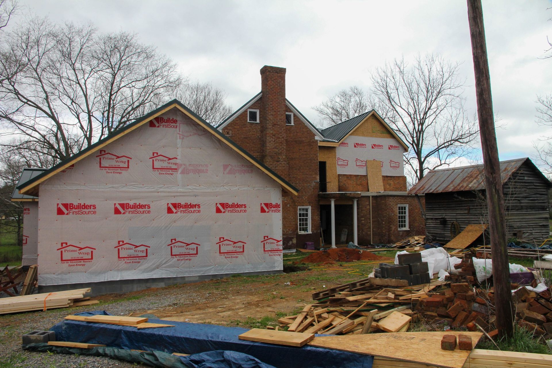 Construction on a brick house with a new wing covered in white protective house wrap, next to an old wooden barn.