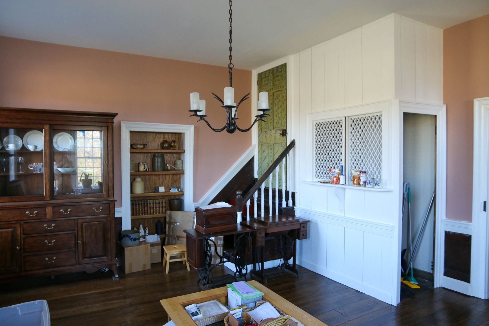 A rustic living space with peach walls, a wooden china cabinet, a staircase, and a wooden sewing desk under a chandelier.