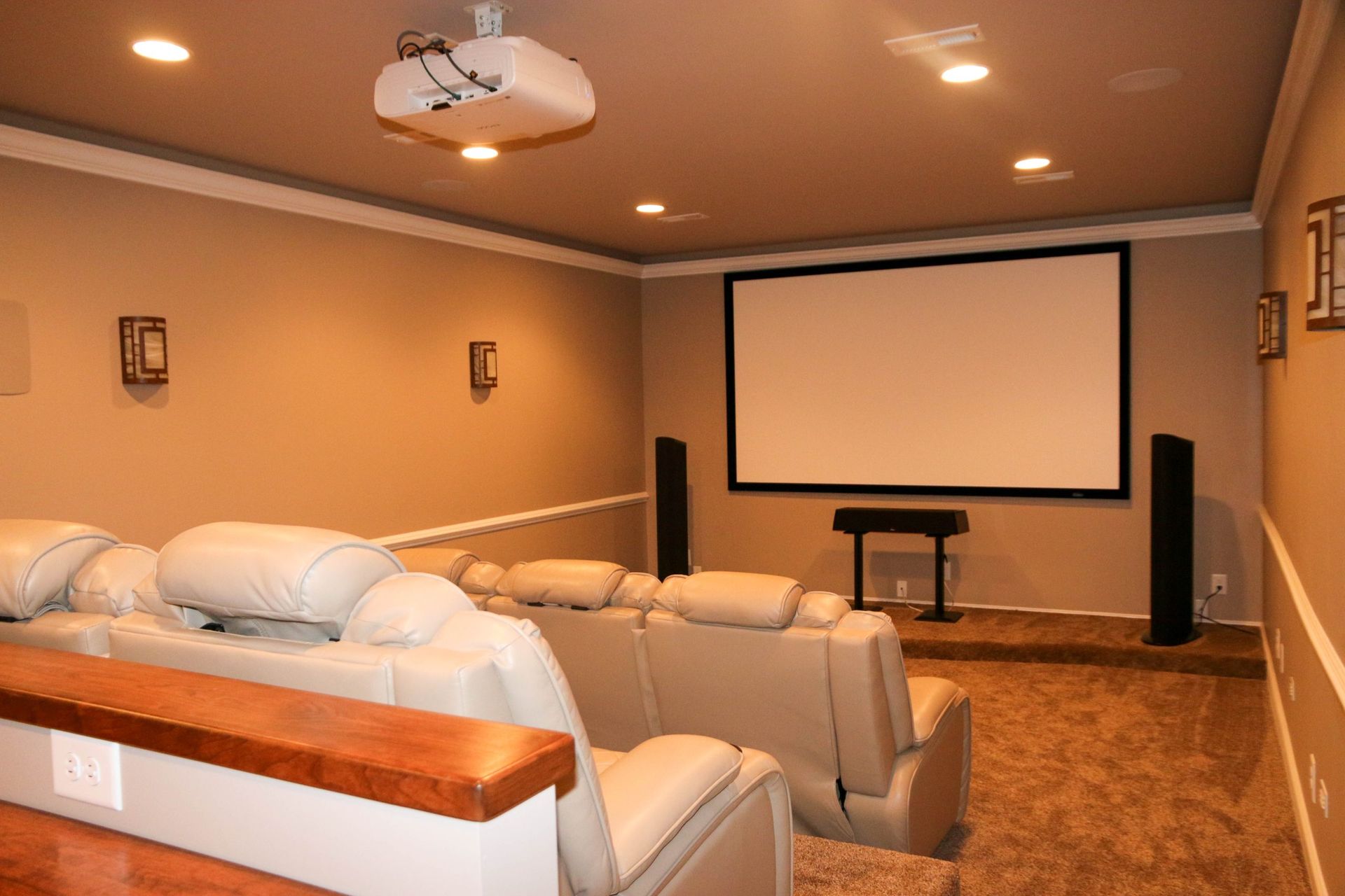 Home theater with beige reclining chairs, a large projection screen, speakers, and a ceiling-mounted projector.