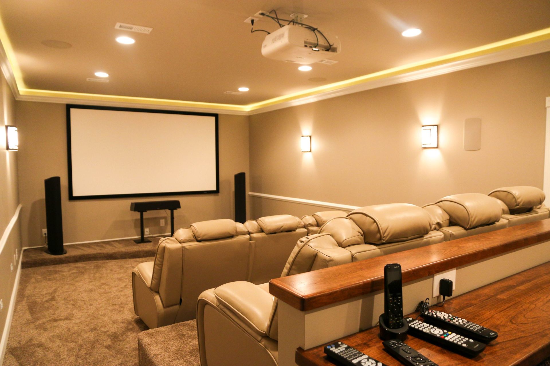 A home theater with a projector, large screen, and two rows of tan leather recliner seats on a carpeted floor.