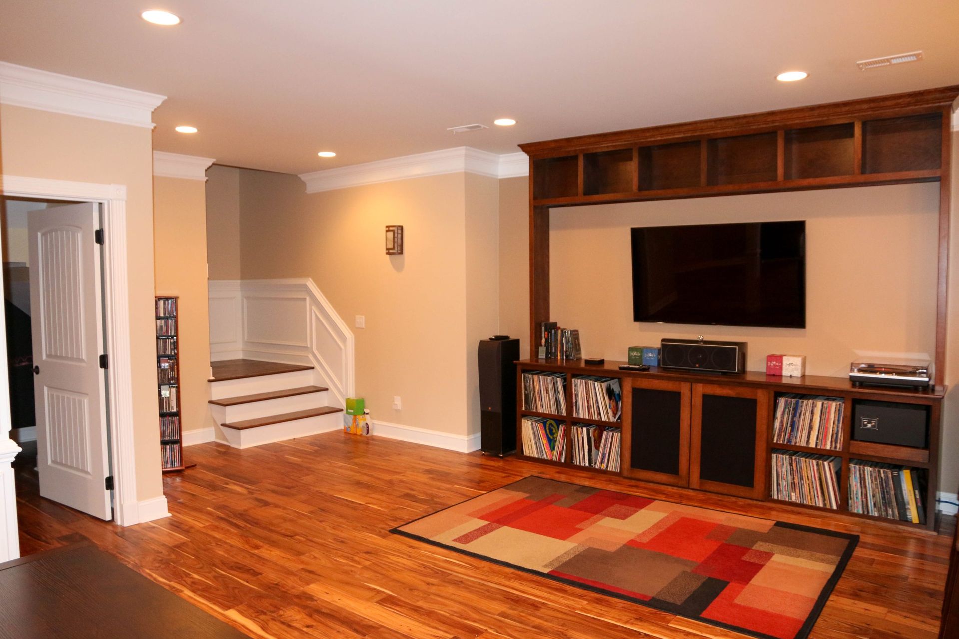 A home media room with wood floors, a large built-in TV unit, a rug with red accents, and stairs leading to an upper level.
