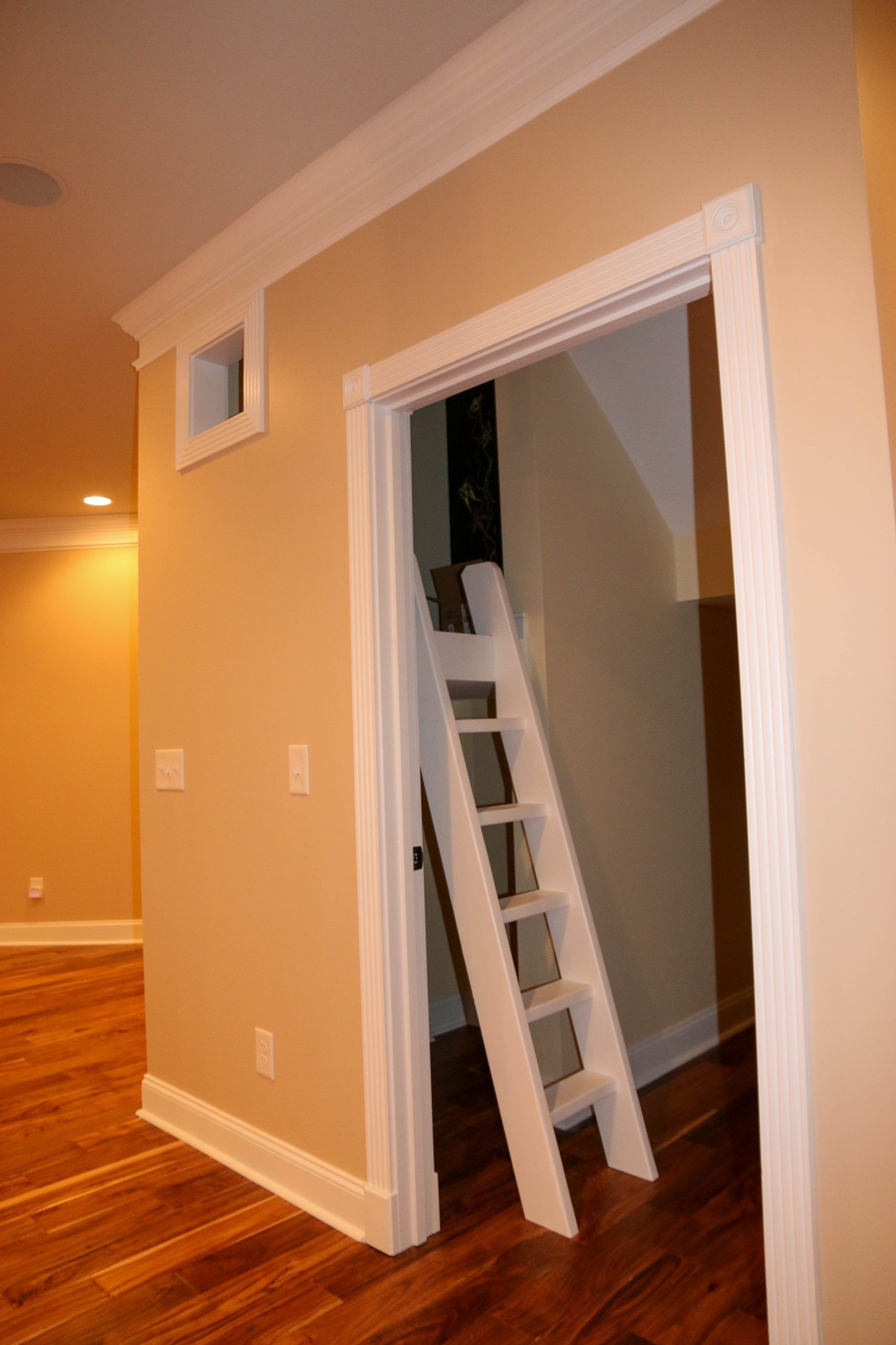 A white wooden ladder inside a doorway leading to an unlit, enclosed space within a room with tan walls and wood floors.