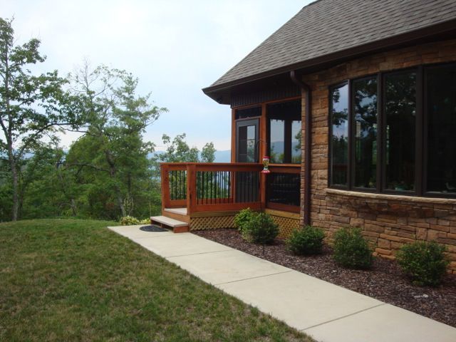 A stone-walled house with a wooden deck and screened porch overlooks a forested mountain landscape.