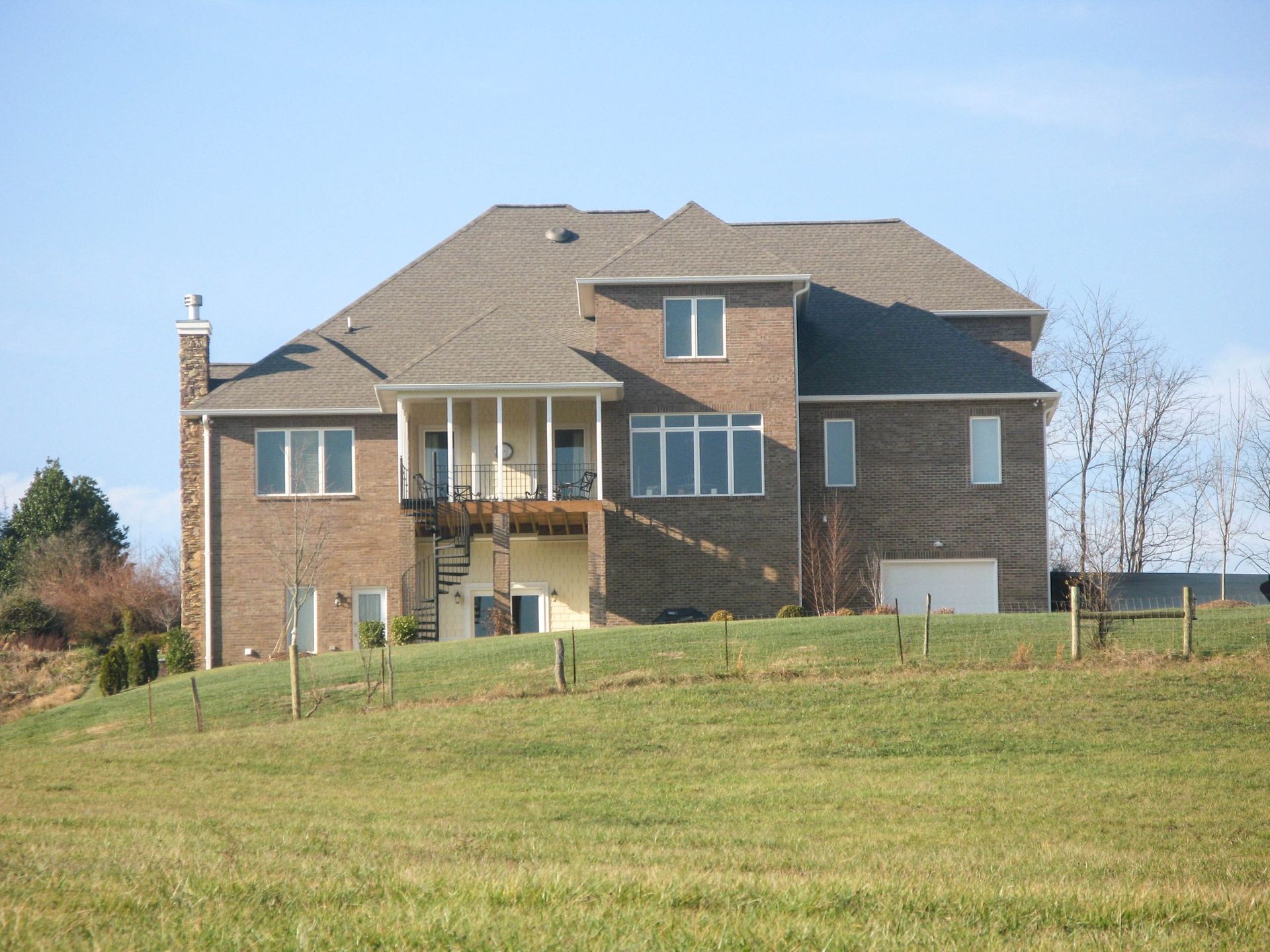 A two-story brick house with a brown shingled roof, a back deck, a spiral staircase, and a large grassy lawn.