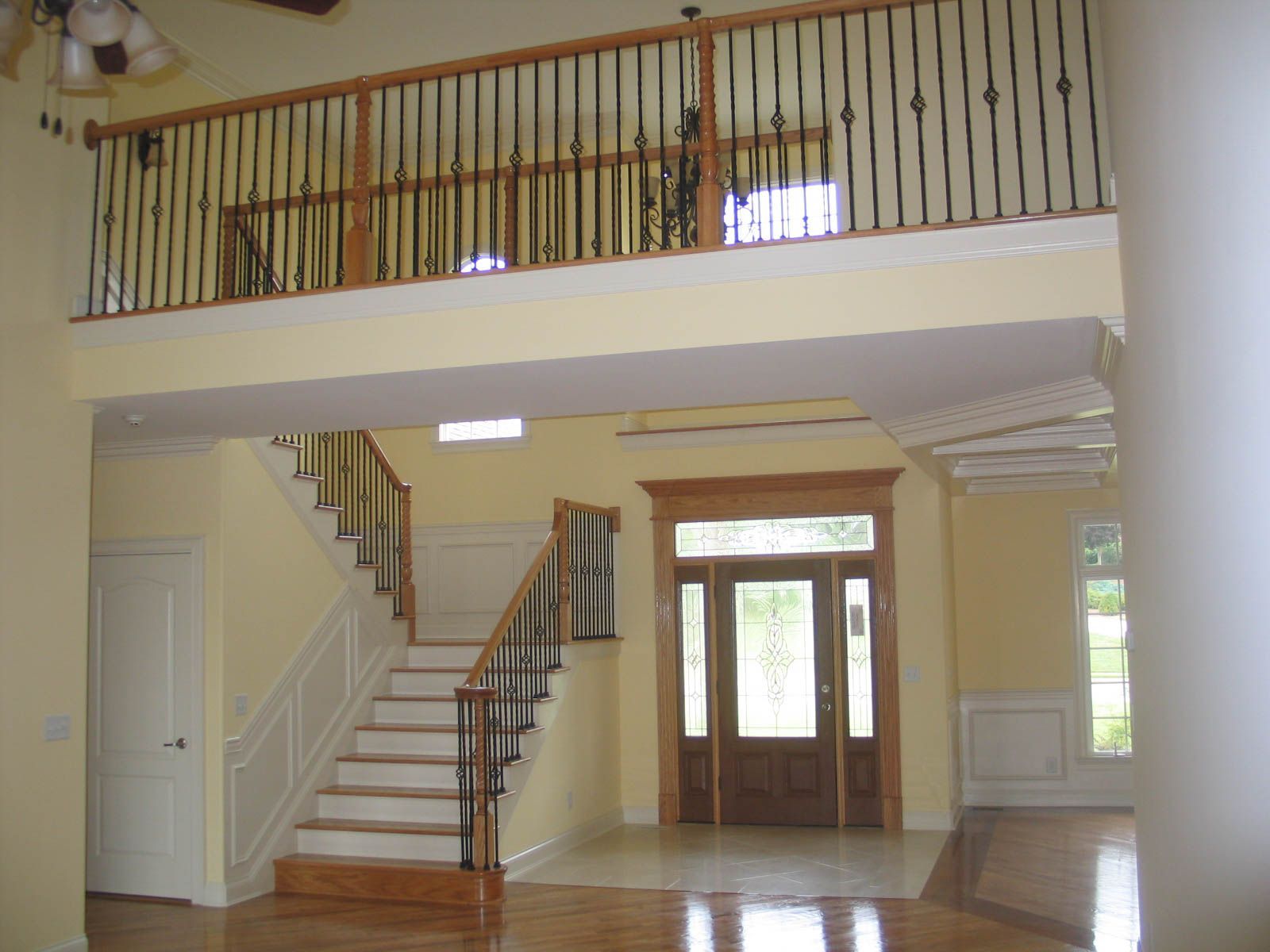 A two-story foyer with yellow walls, a wooden staircase, a balcony with black metal railings, and a wooden front door.