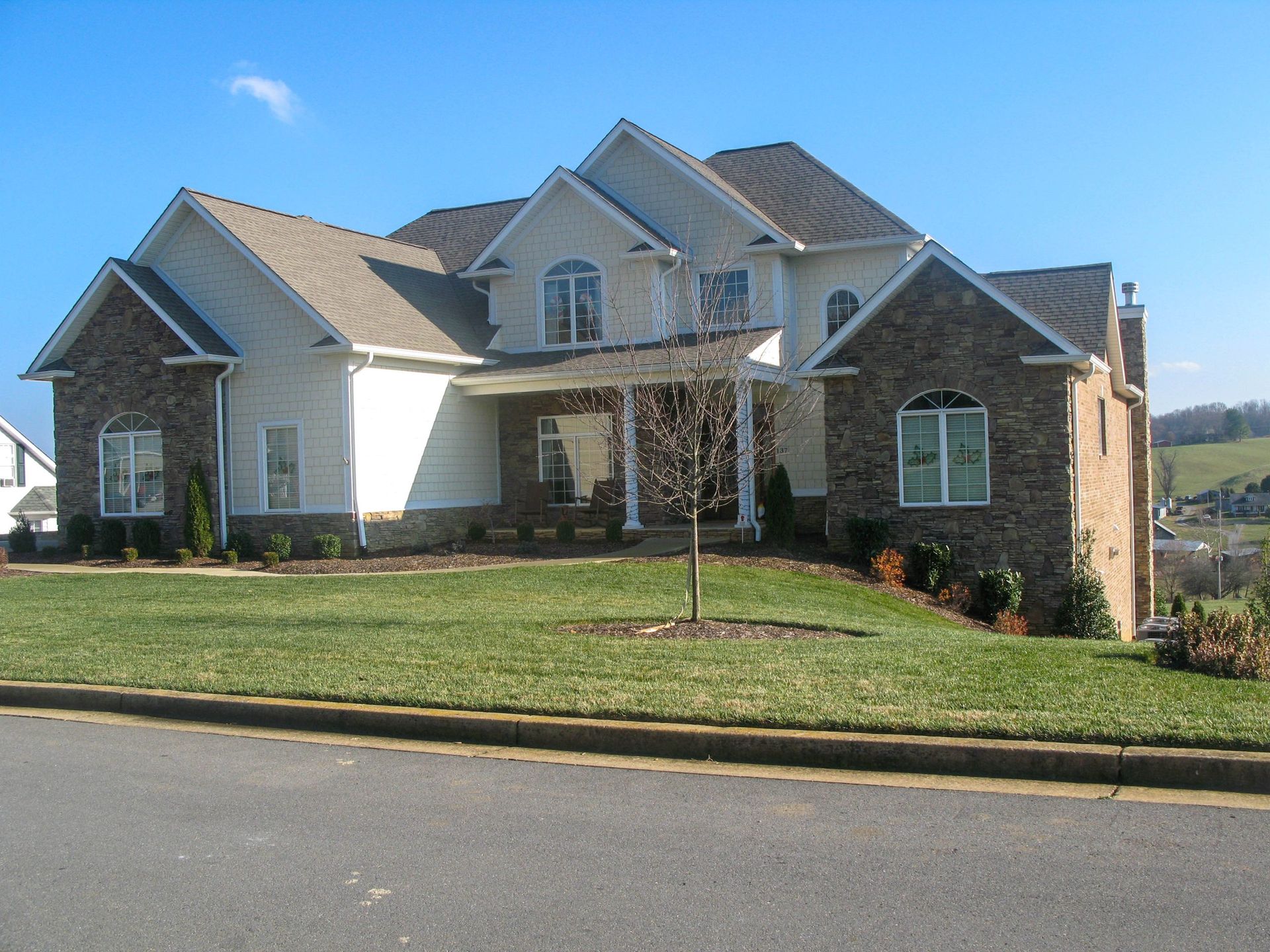 A two-story suburban house with stone and cream siding, a dark roof, and a front yard, under a clear blue sky.