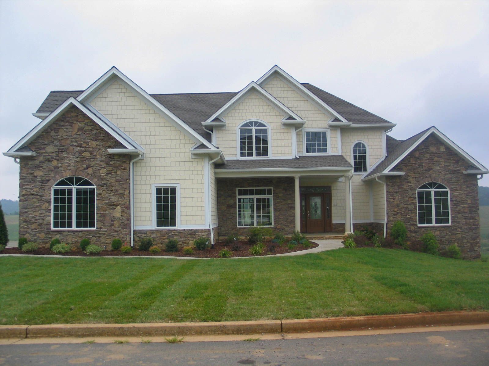 A two-story suburban house with beige siding, stone facade, and a dark roof, set on a grassy lot under an overcast sky.