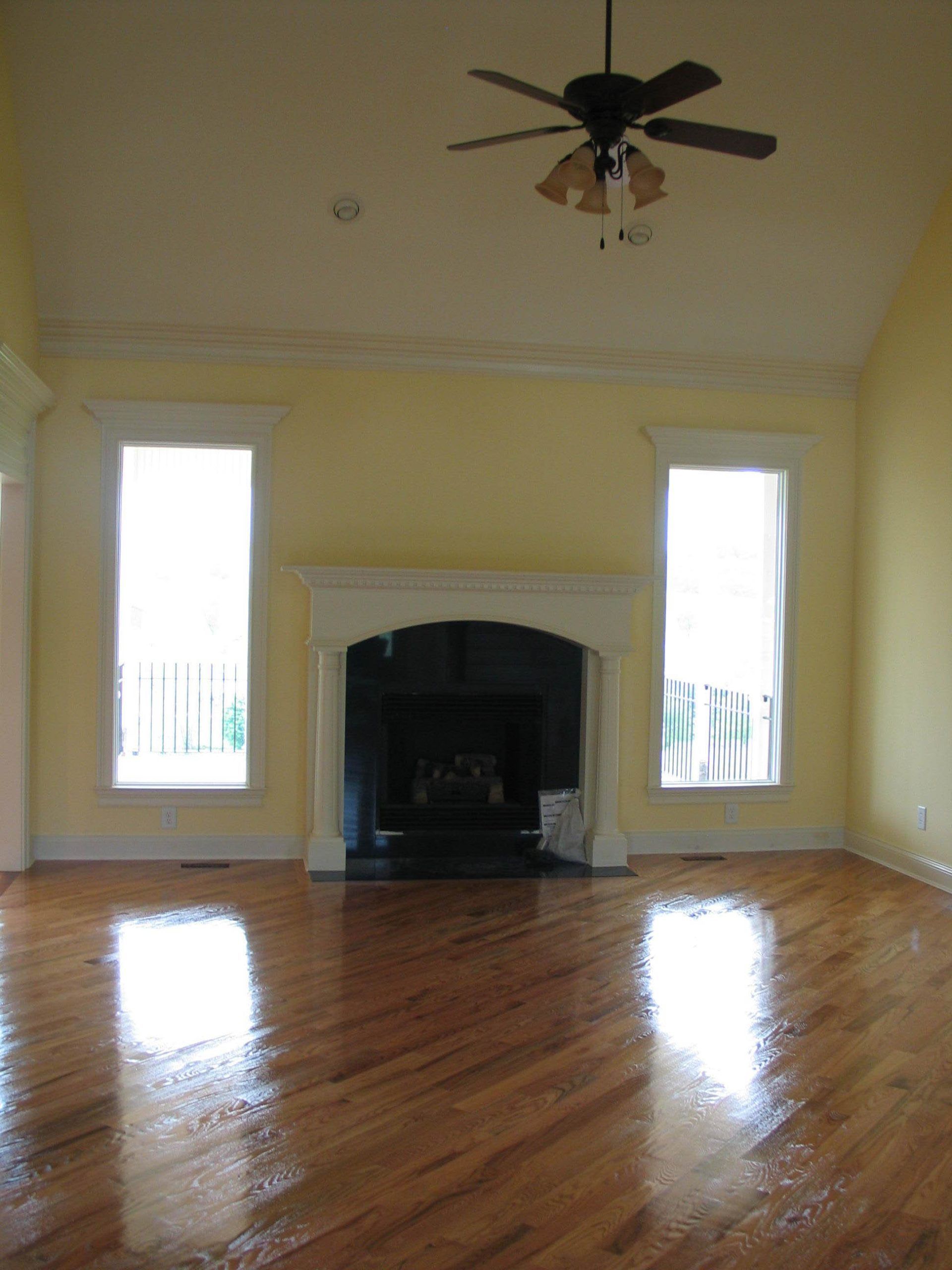 A yellow living room with hardwood floors, a central fireplace, two tall windows, and a ceiling fan.