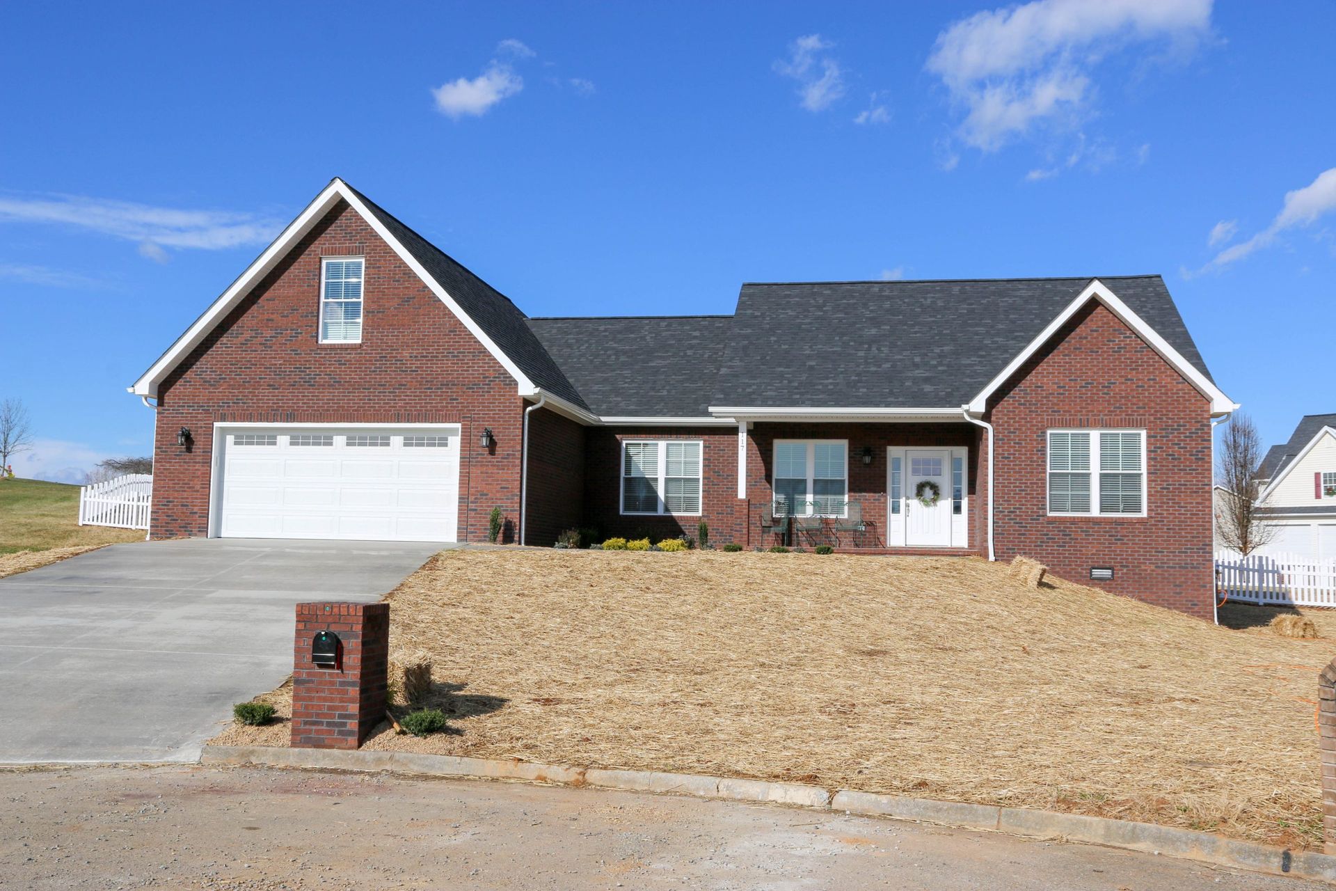 A single-story brick house with a two-car garage, white trim, and a freshly mulched front yard under a blue sky.