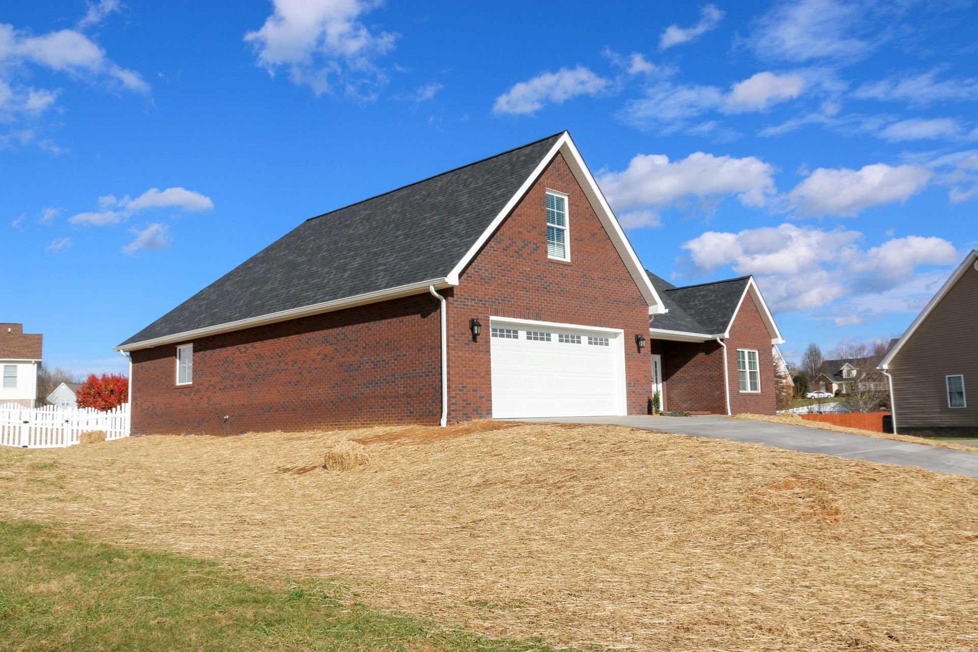 A newly constructed red brick house with a two-car garage, dark shingled roof, and unfinished yard under a blue sky.