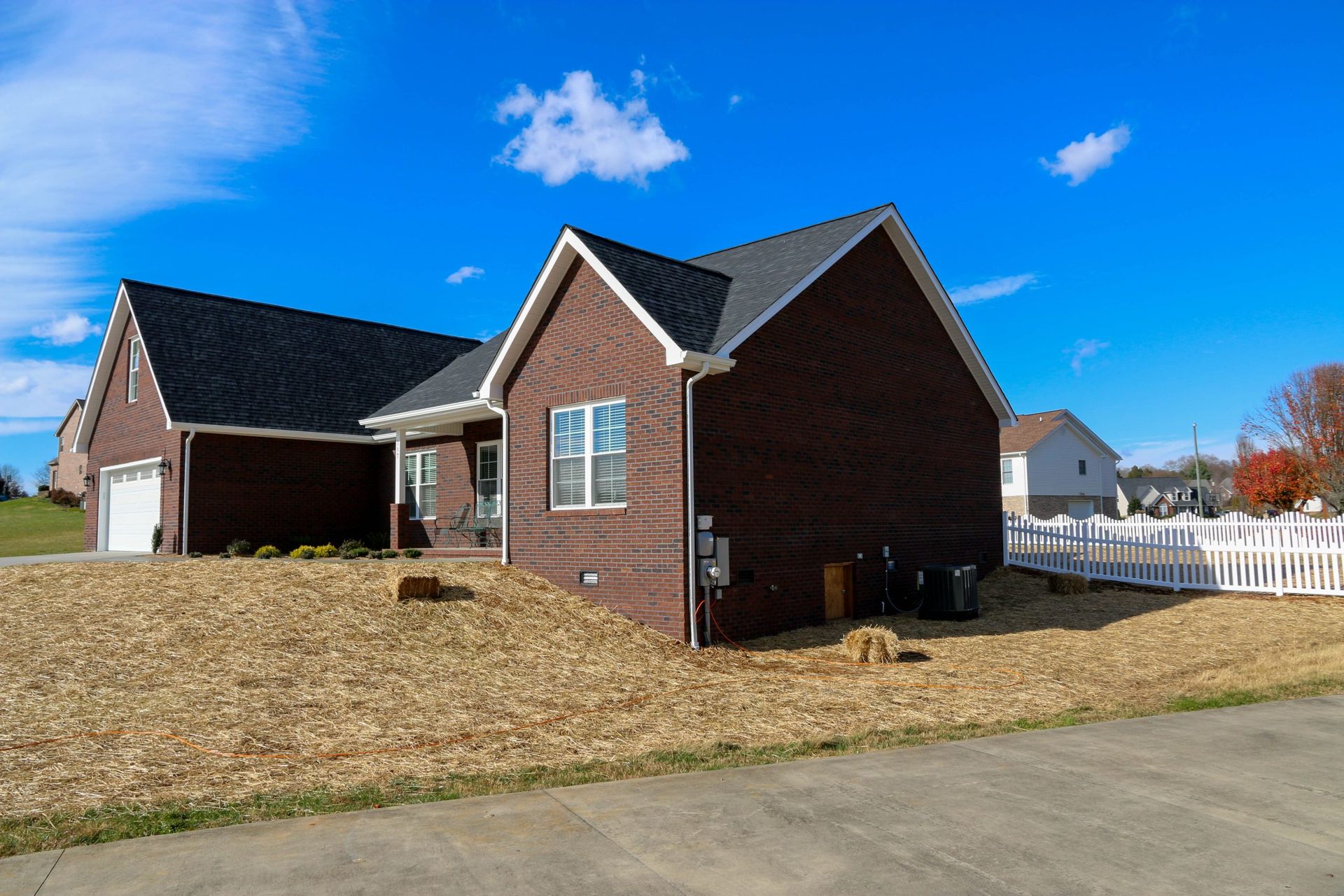 A brick suburban house with a dark shingled roof, a garage, and a white picket fence on a sunny day.