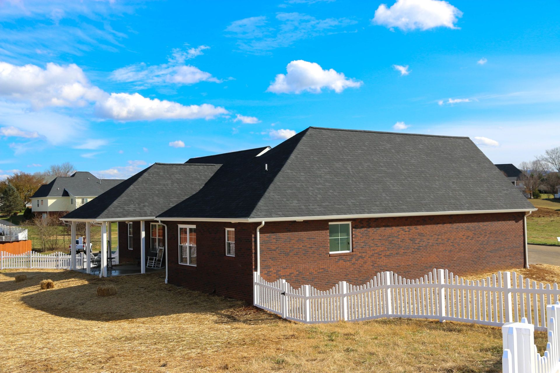 A brick house with a dark shingled roof under a blue sky with white clouds, surrounded by a white picket fence.