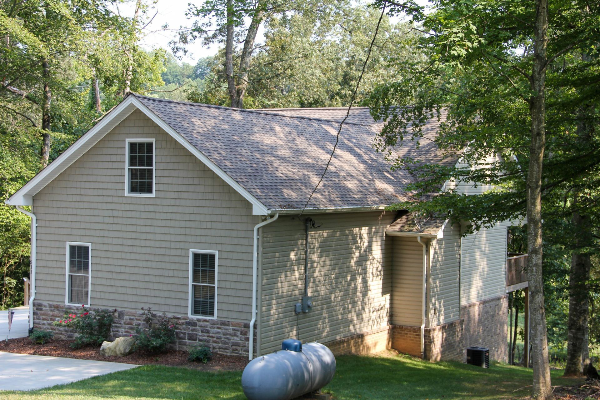 A two-story, tan-sided house with stone veneer base and a shingled roof, nestled in a wooded area with a propane tank.