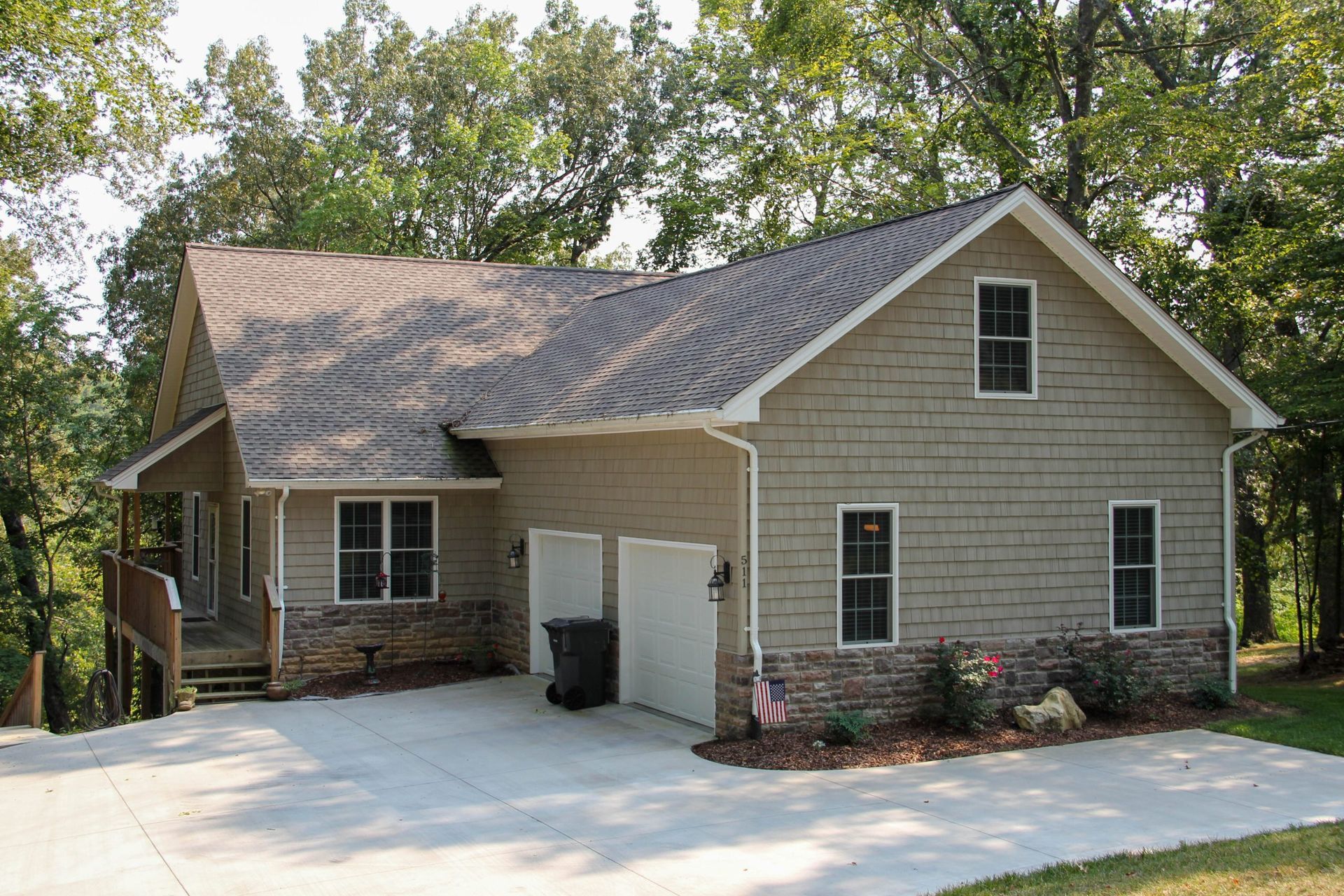 A tan house with stone accents, a two-car garage, and a wrap-around porch surrounded by mature trees.