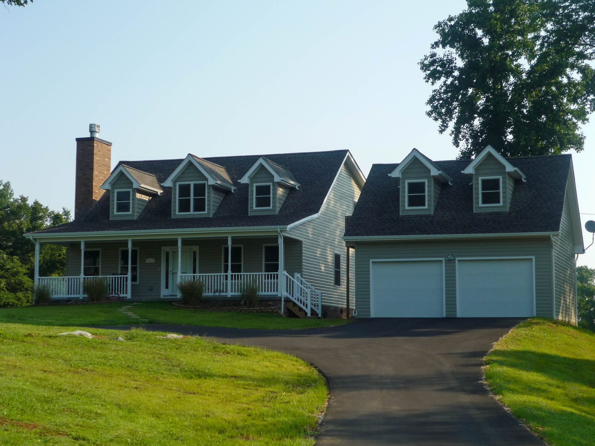 A light-gray, two-story house with a front porch, a brick chimney, and a detached two-car garage on a grassy lot.