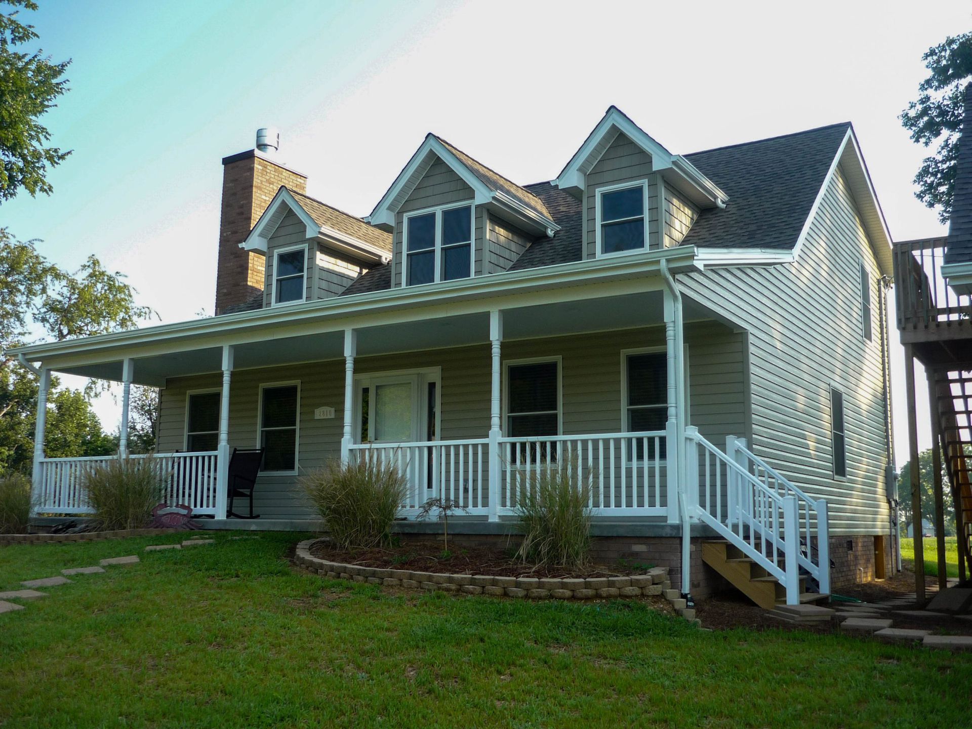 A two-story tan house with a large front porch, white railings, two dormer windows, and a brick chimney.