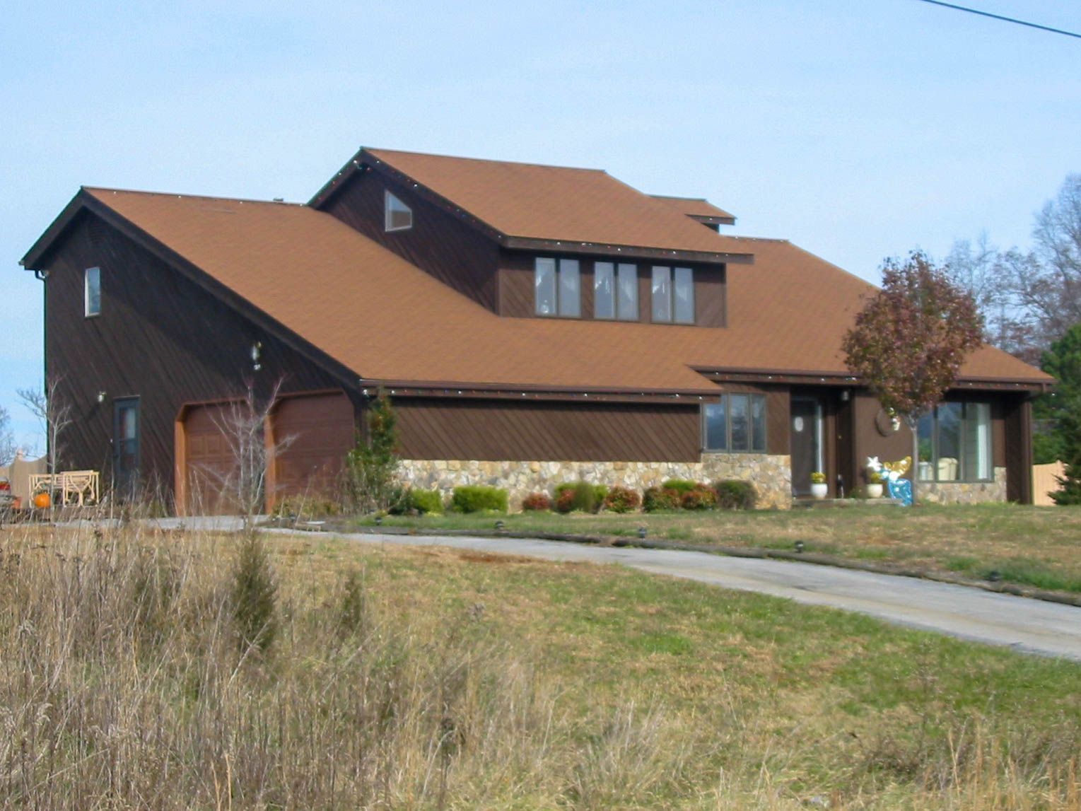 A brown, multi-level house with dark siding, a stone base, and a driveway set in a rural, grassy landscape.