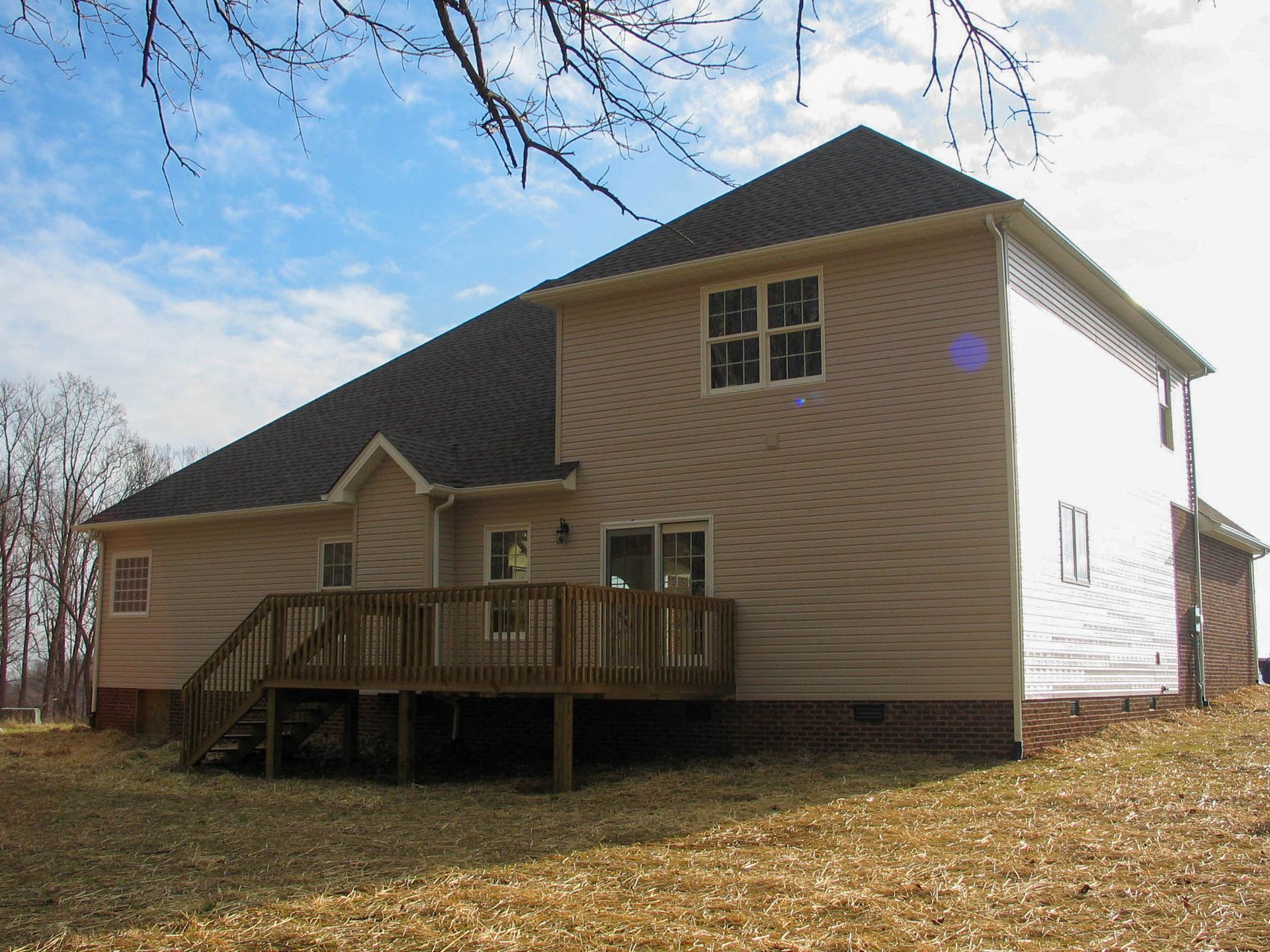 A two-story tan house with a dark roof and a wooden deck, set against a bright, cloudy sky on a grassy lot.