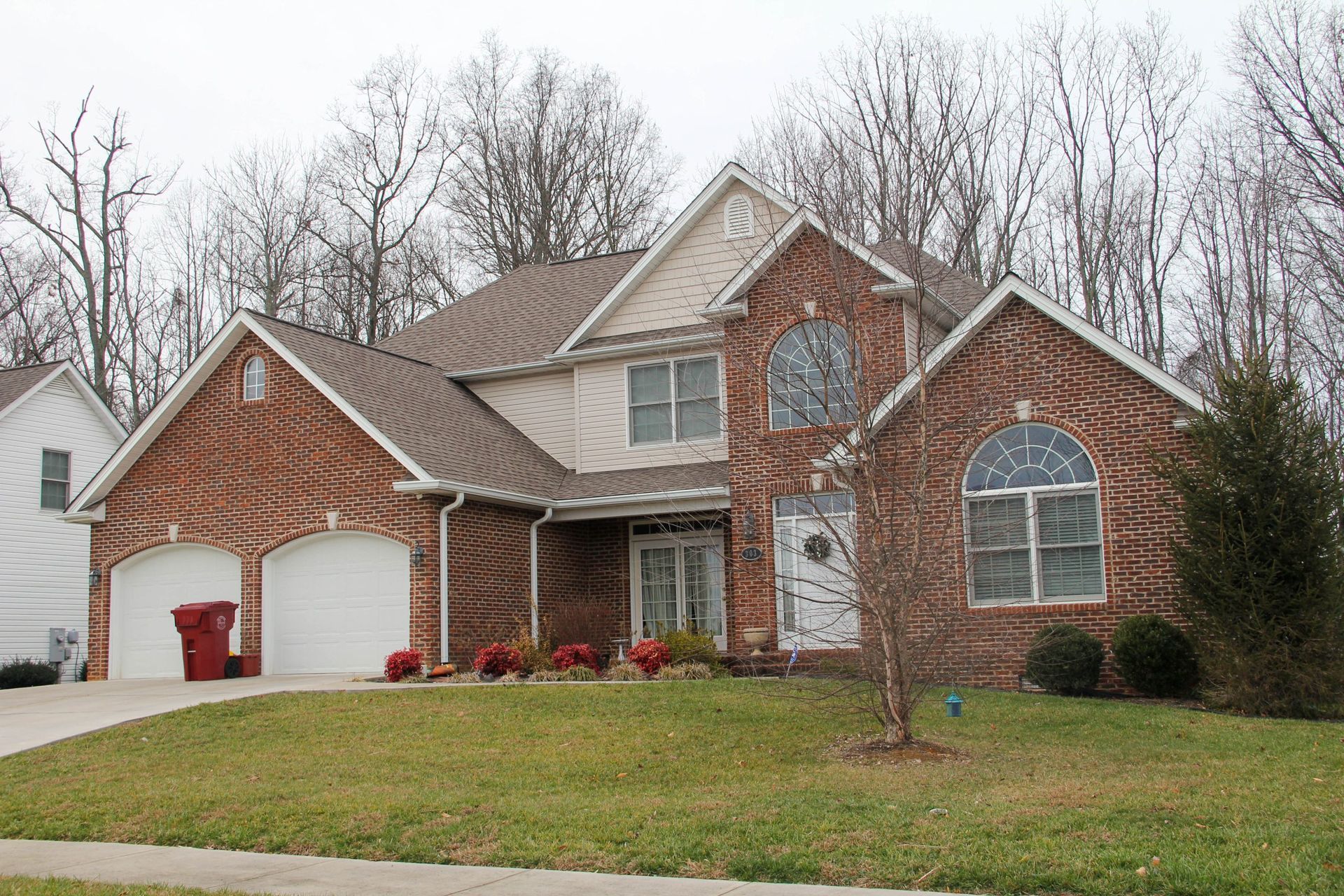 A two-story suburban brick house with a two-car garage and a landscaped front lawn under a cloudy sky.