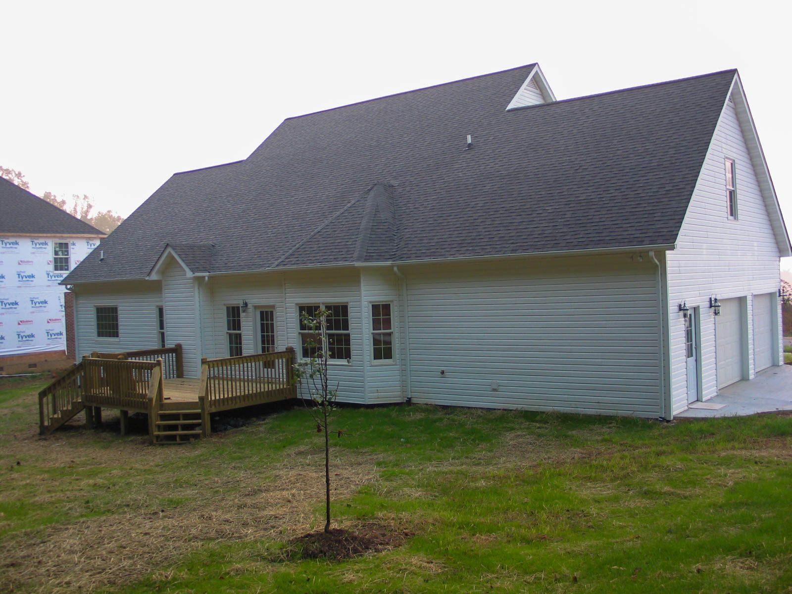 A white suburban house with a dark shingled roof, a wooden back deck, and a multi-car garage on a grassy lot.
