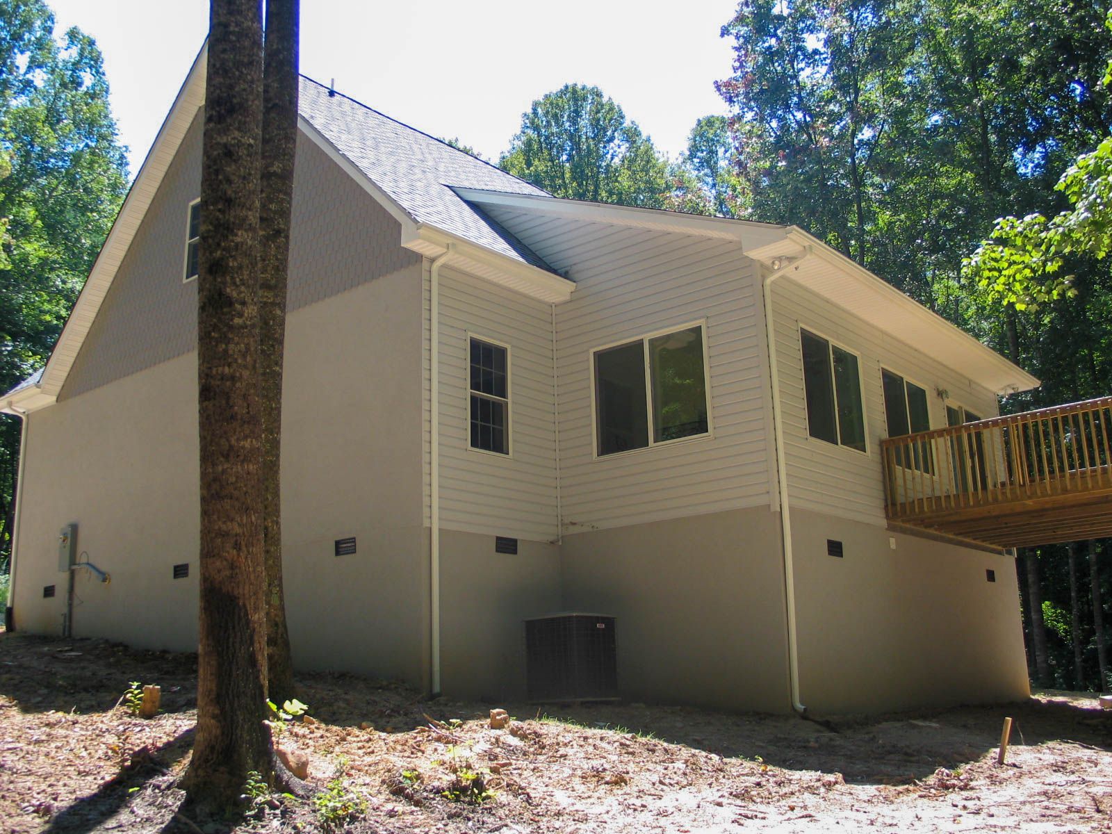 Exterior of a beige-sided house with a raised wooden deck, surrounded by trees on a sloped, wooded lot.
