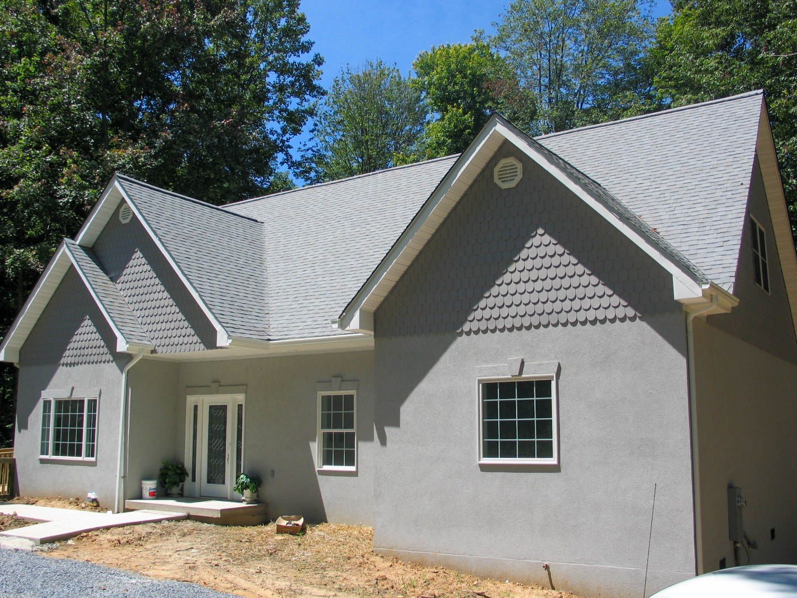 A single-story gray house with a peaked roof, decorative siding accents, and a front entry, set against a tree line.