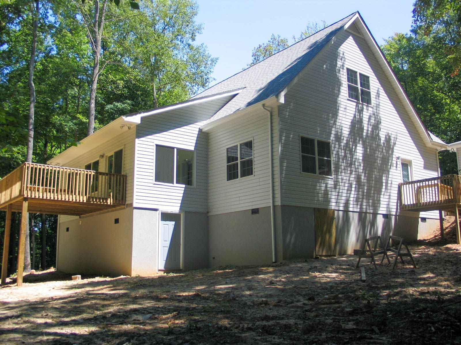 A two-story house with light siding and a gray foundation, featuring two wooden decks surrounded by a wooded area.