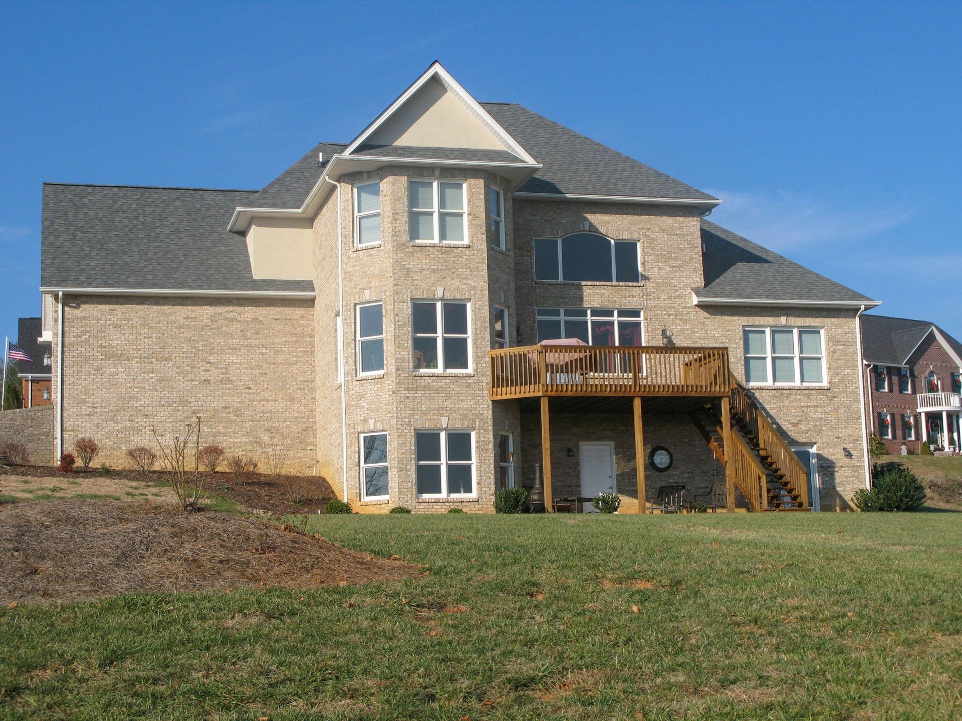 A beige, multi-story brick suburban house with a wooden deck and staircase, set against a clear blue sky.