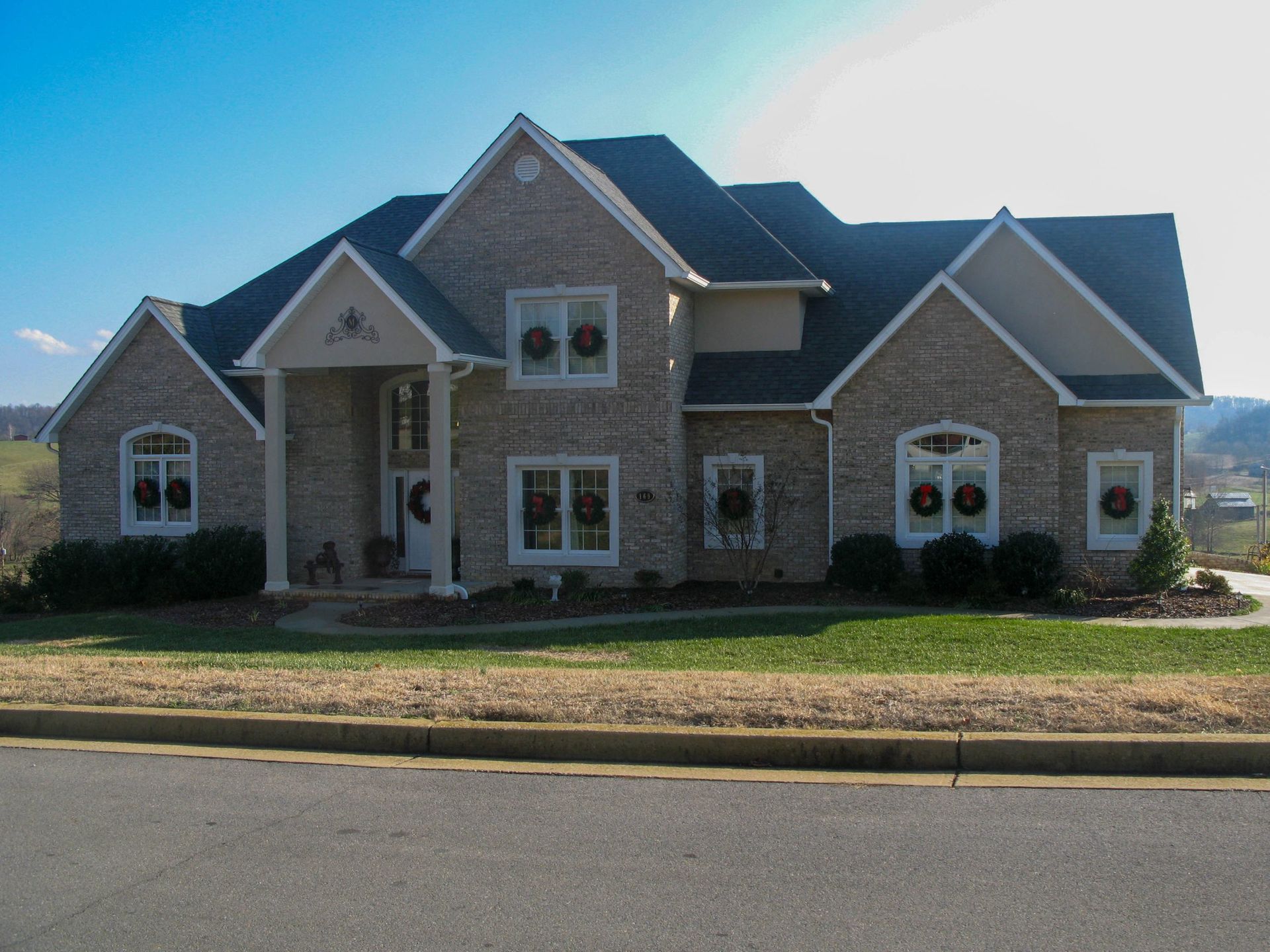 A large, two-story tan brick house with a dark roof and decorative holiday wreaths on its front-facing windows.