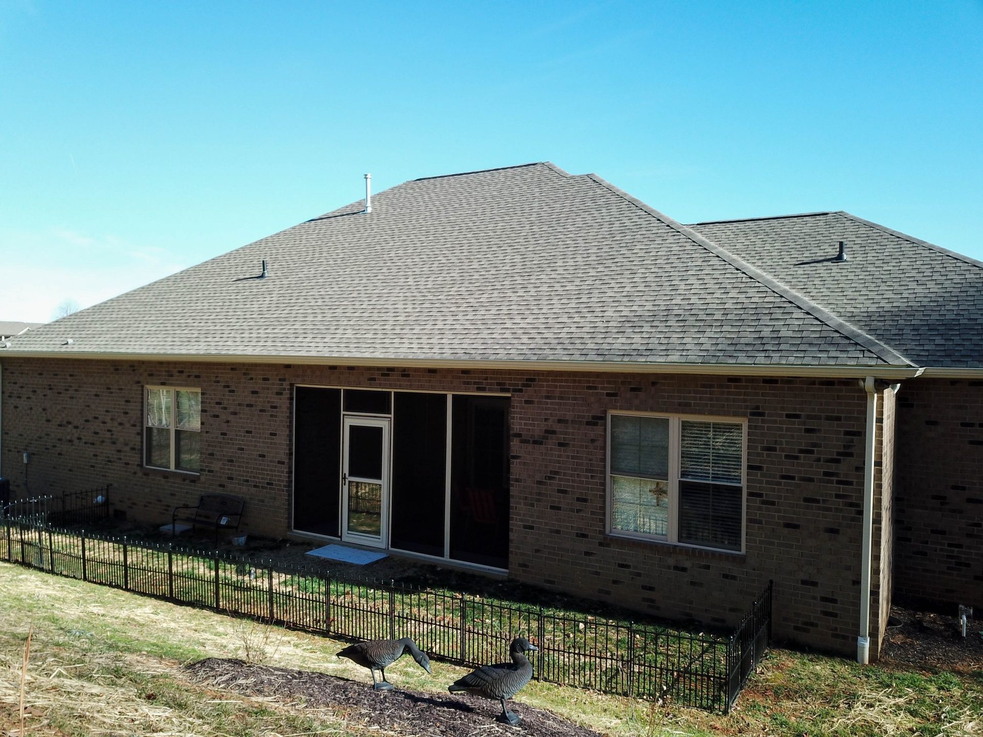 A brick house with a dark shingled roof, screened porch, and a metal fence in the backyard under a clear blue sky.