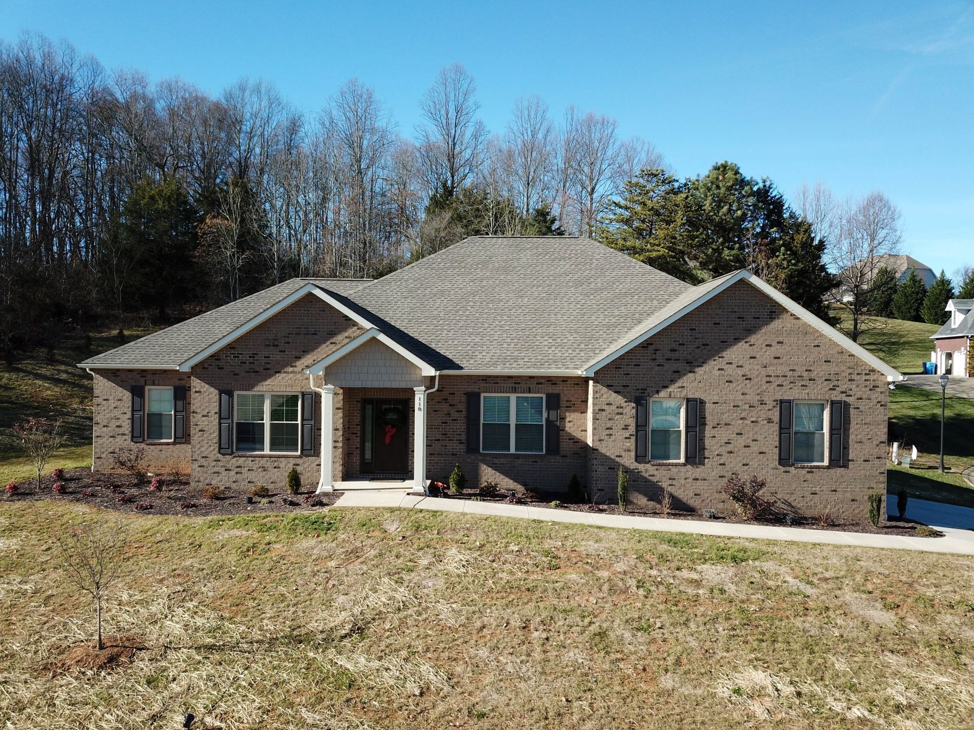 A single-story, brown brick house with a dark gray shingled roof, centered on a grassy lot under a clear blue sky.