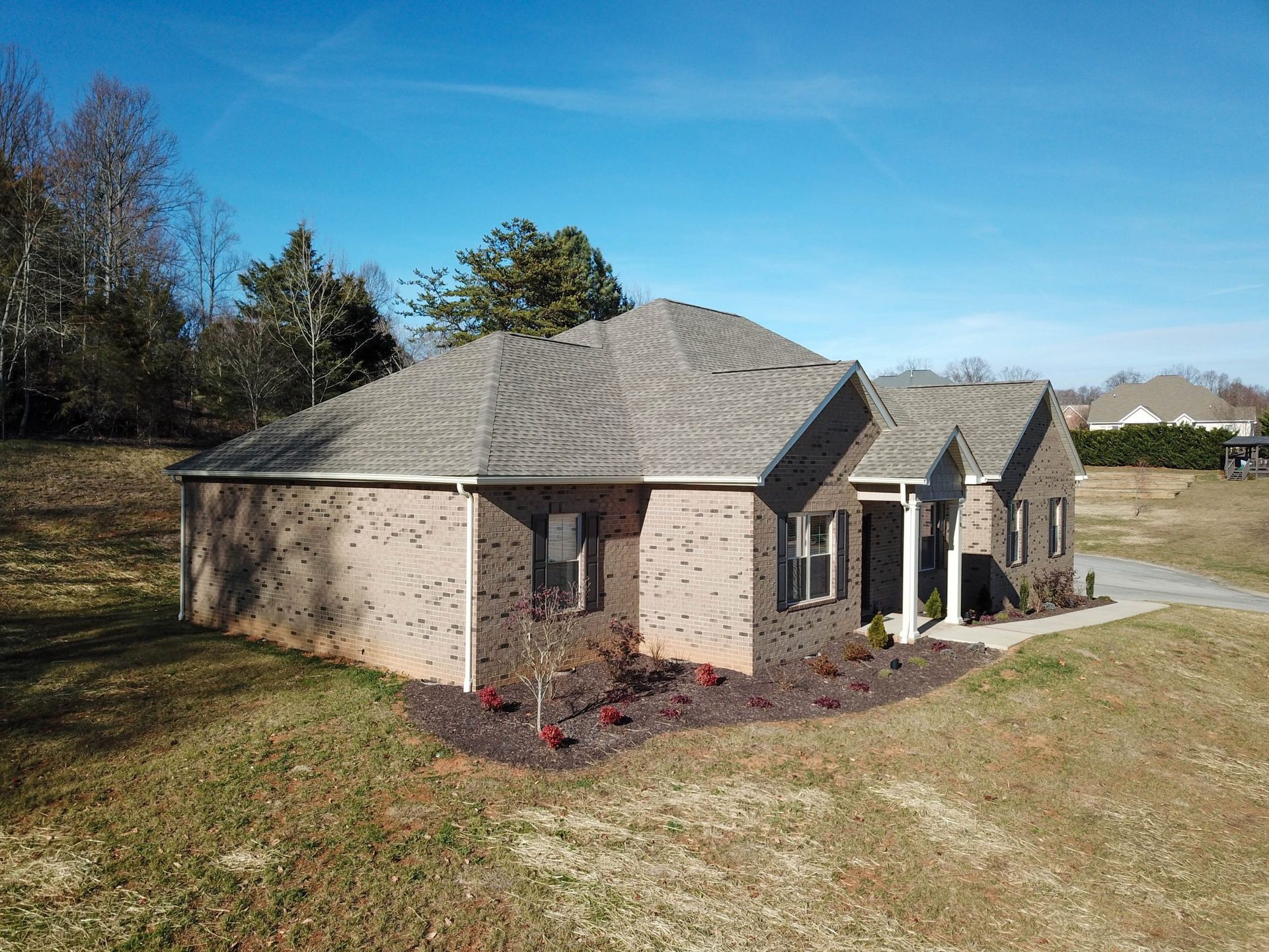 A one-story brick house with a gray shingled roof, surrounded by a grassy lawn under a clear blue sky.