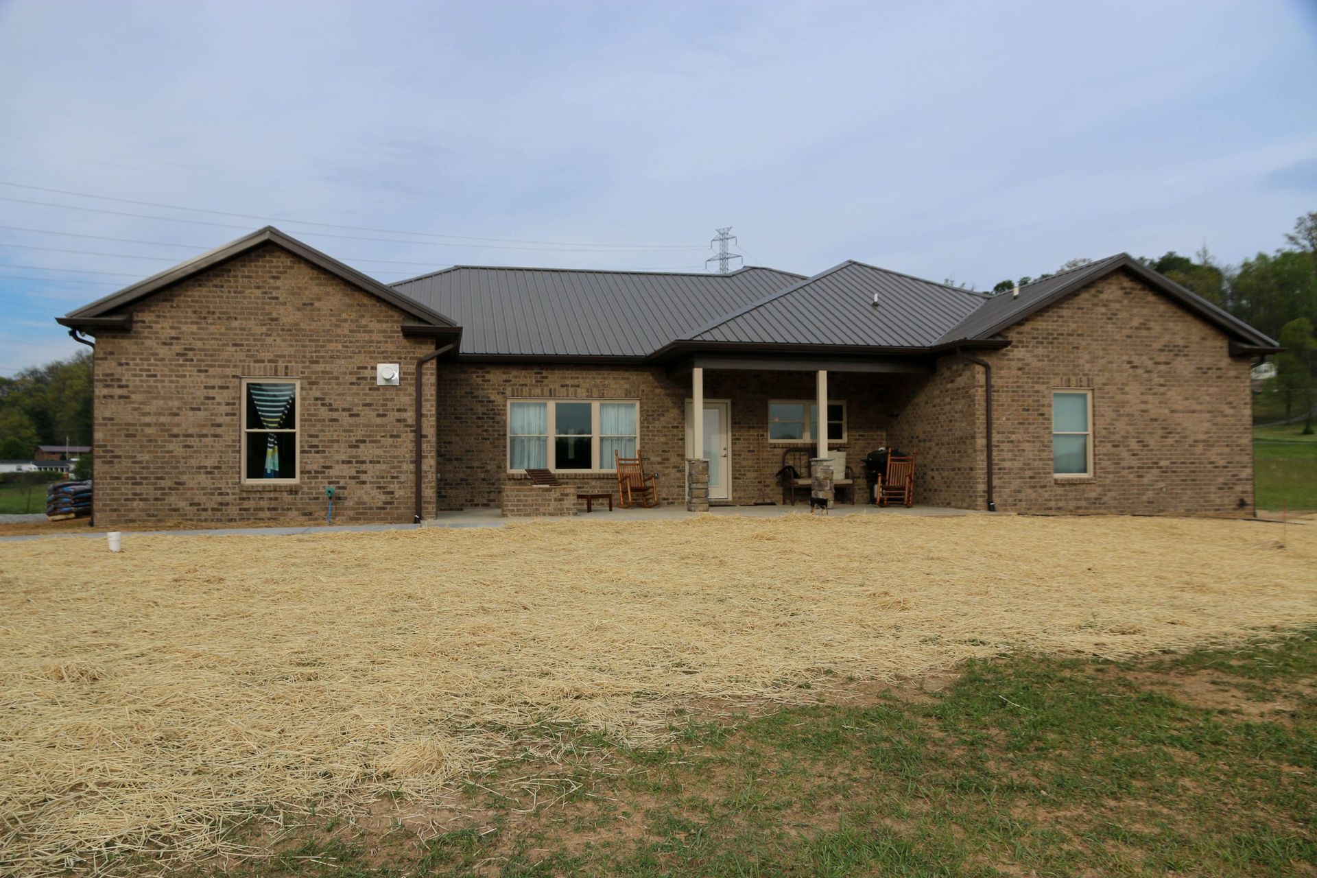 A single-story, brown brick house with a dark roof and a central covered patio sits on a lot covered in straw mulch.