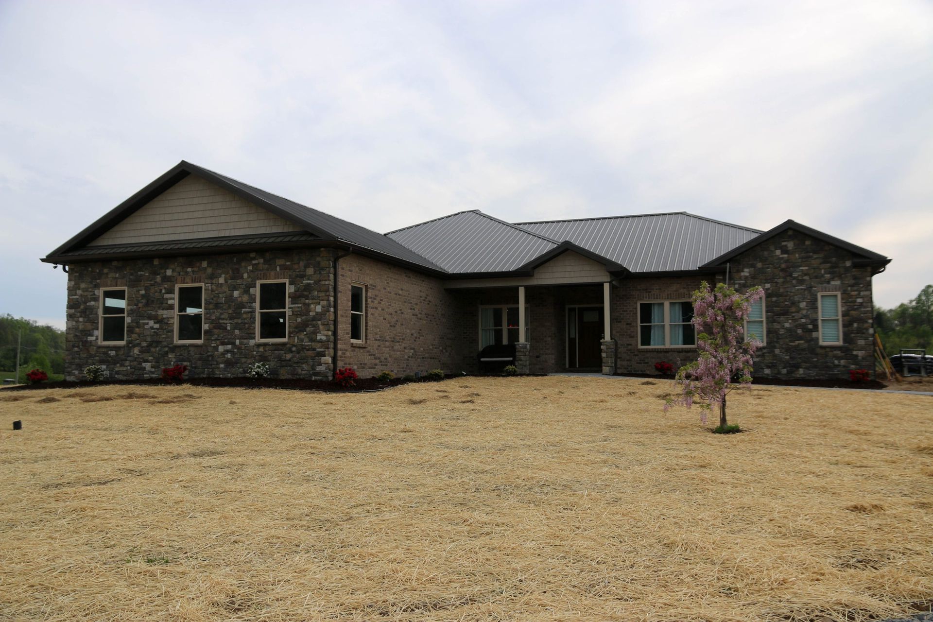 A single-story, stone-veneer house with a dark metal roof, set against a cloudy sky over a field of dry, dormant grass.