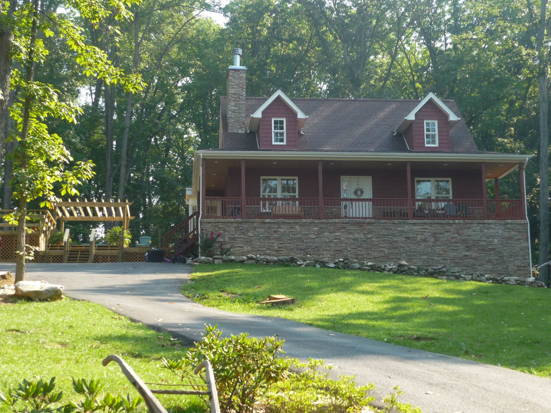 A red, two-story house with a stone base, front porch, and a chimney, situated in a wooded area with a paved driveway.