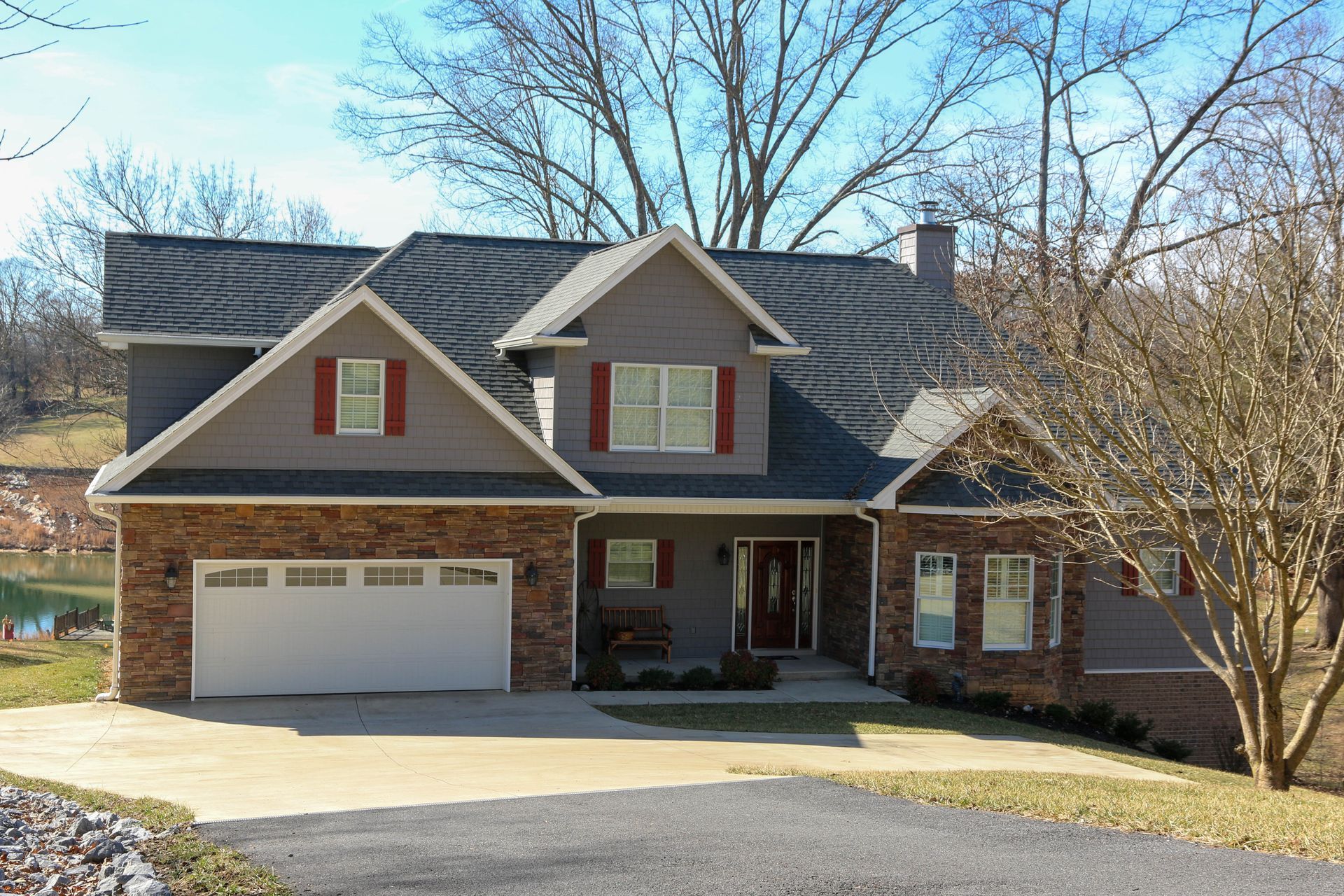 A two-story house with a stone and gray siding exterior, a white garage, red shutters, and trees in a grassy yard.