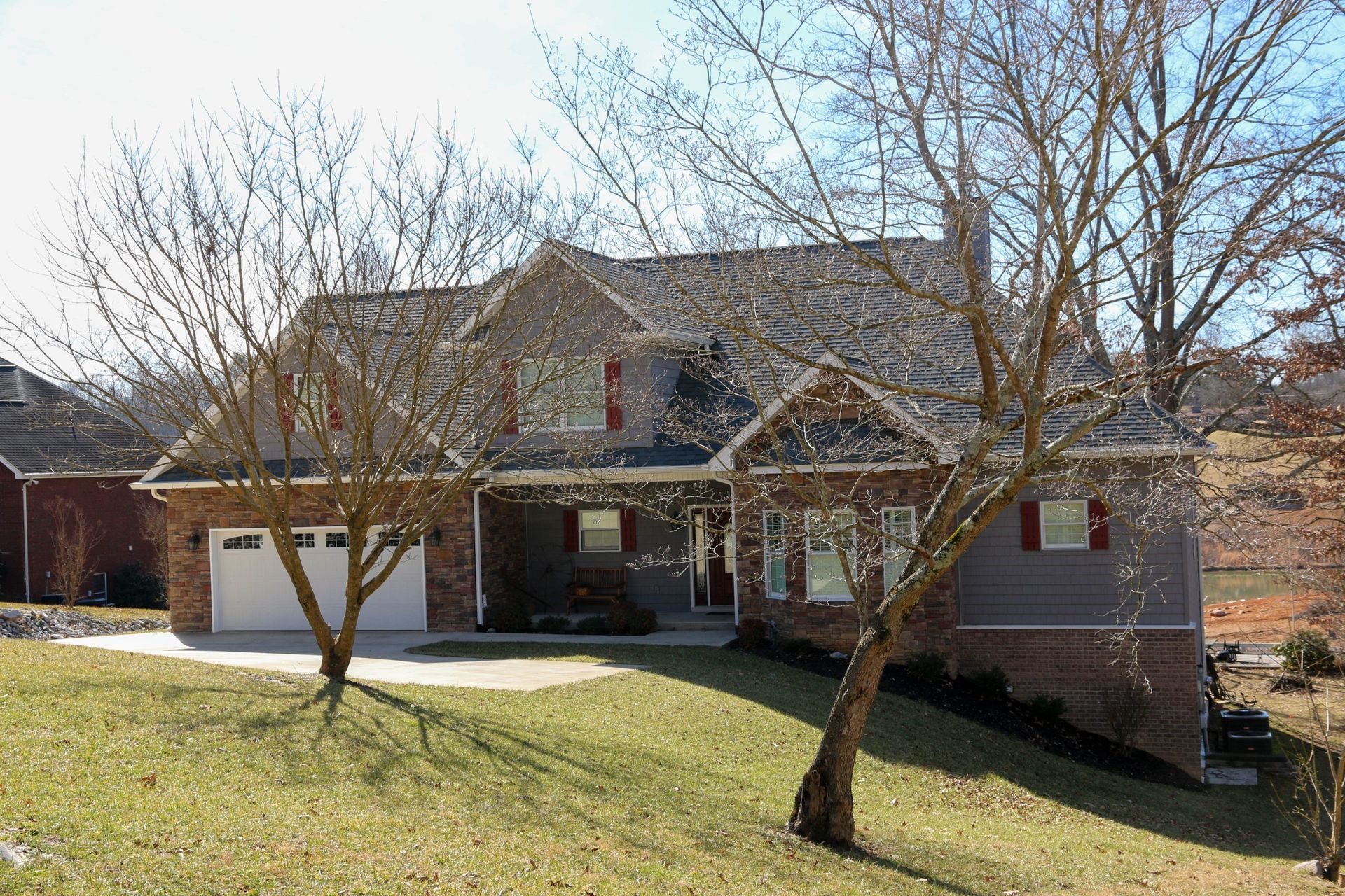 A two-story suburban house with brick and gray siding, a garage, and bare trees in the front yard on a sunny day.