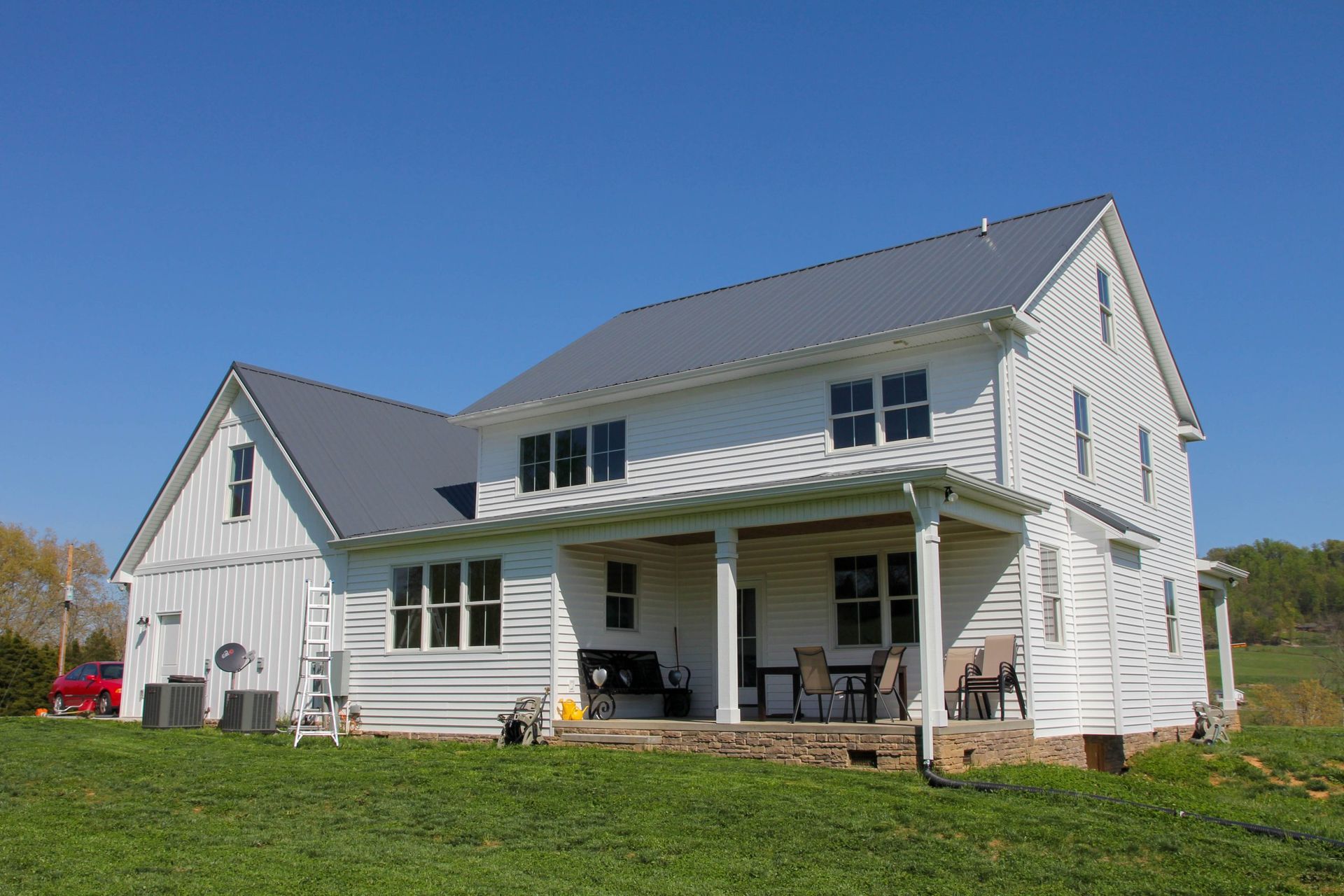 A two-story white farmhouse with a dark metal roof, a covered porch, and a yard under a clear blue sky.