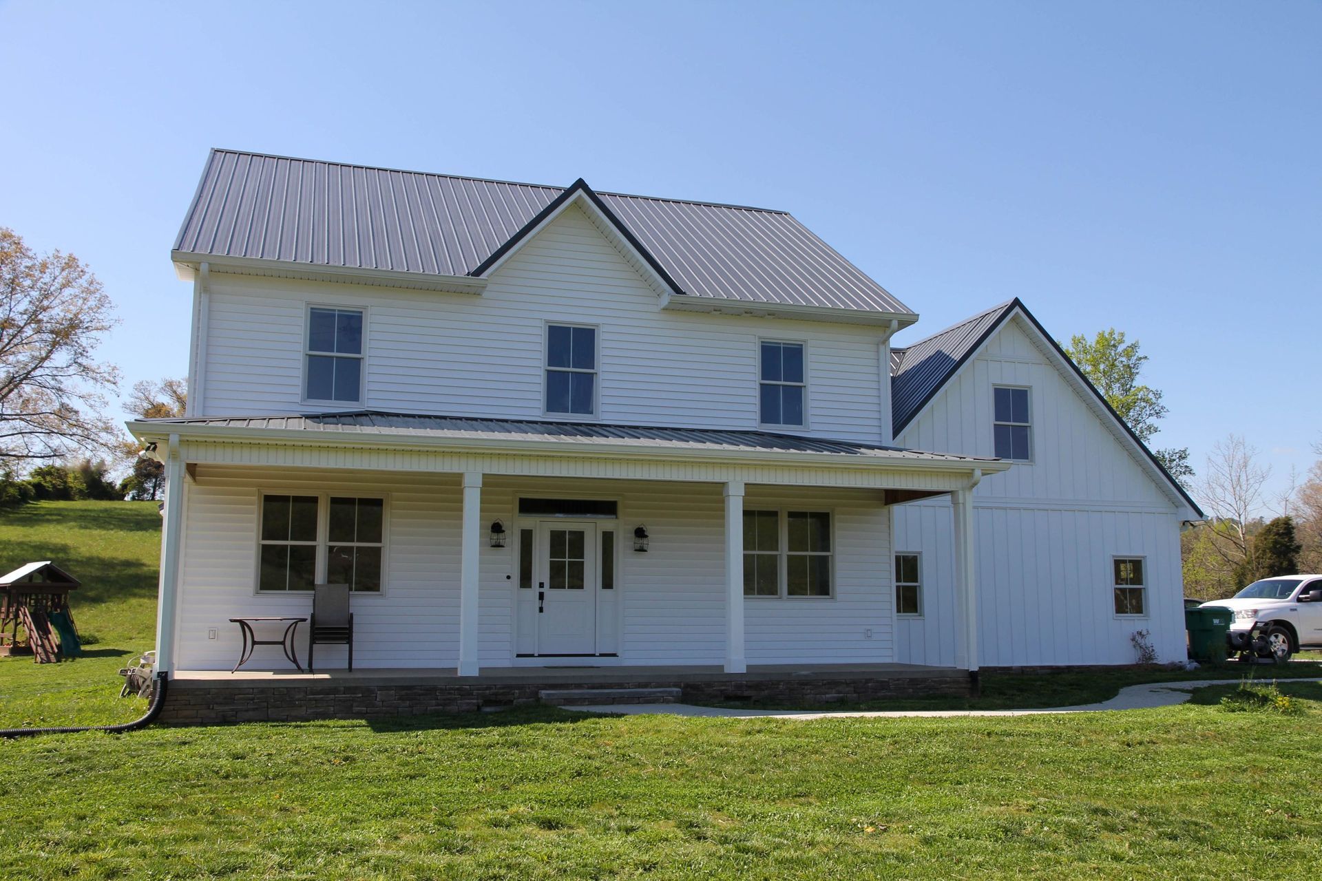 A two-story white farmhouse with a dark metal roof, front porch, and a yard under a clear blue sky.