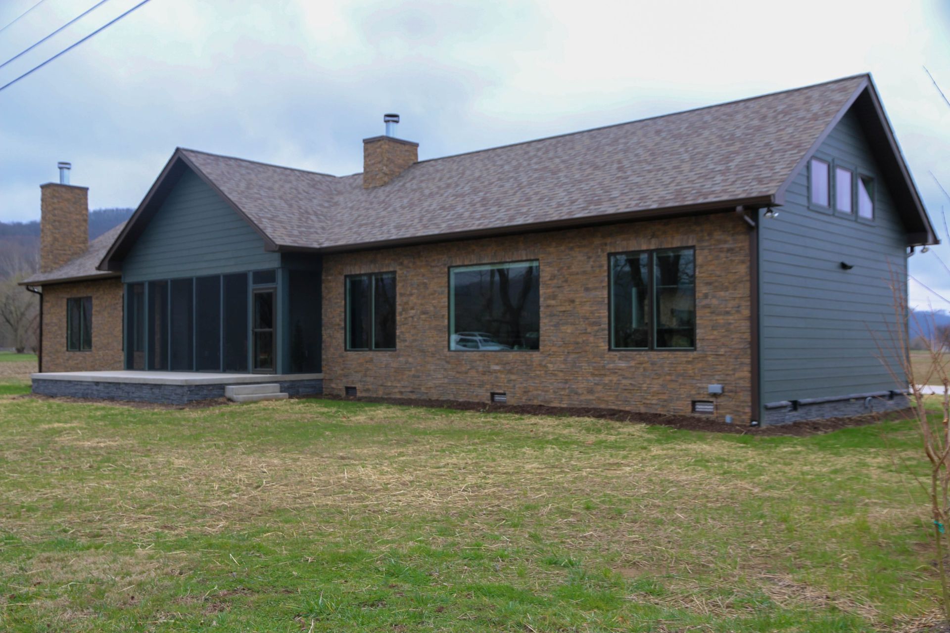 A single-story house with dark green siding, stone veneer, two chimneys, and a screened-in porch on a grassy field.