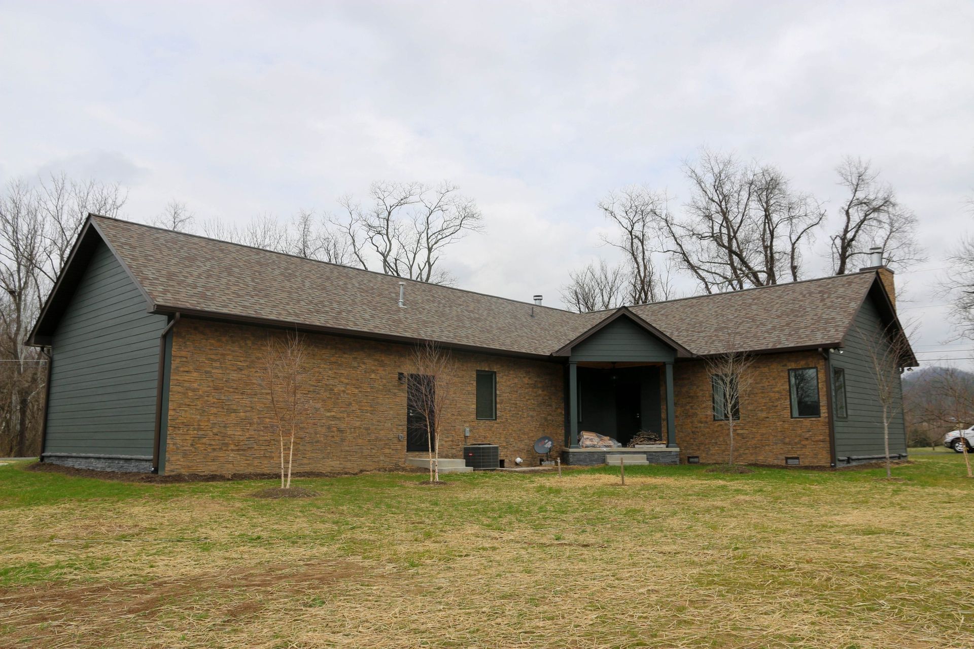 A one-story house with dark gray siding and a tan brick facade, featuring a central porch set against a cloudy sky.