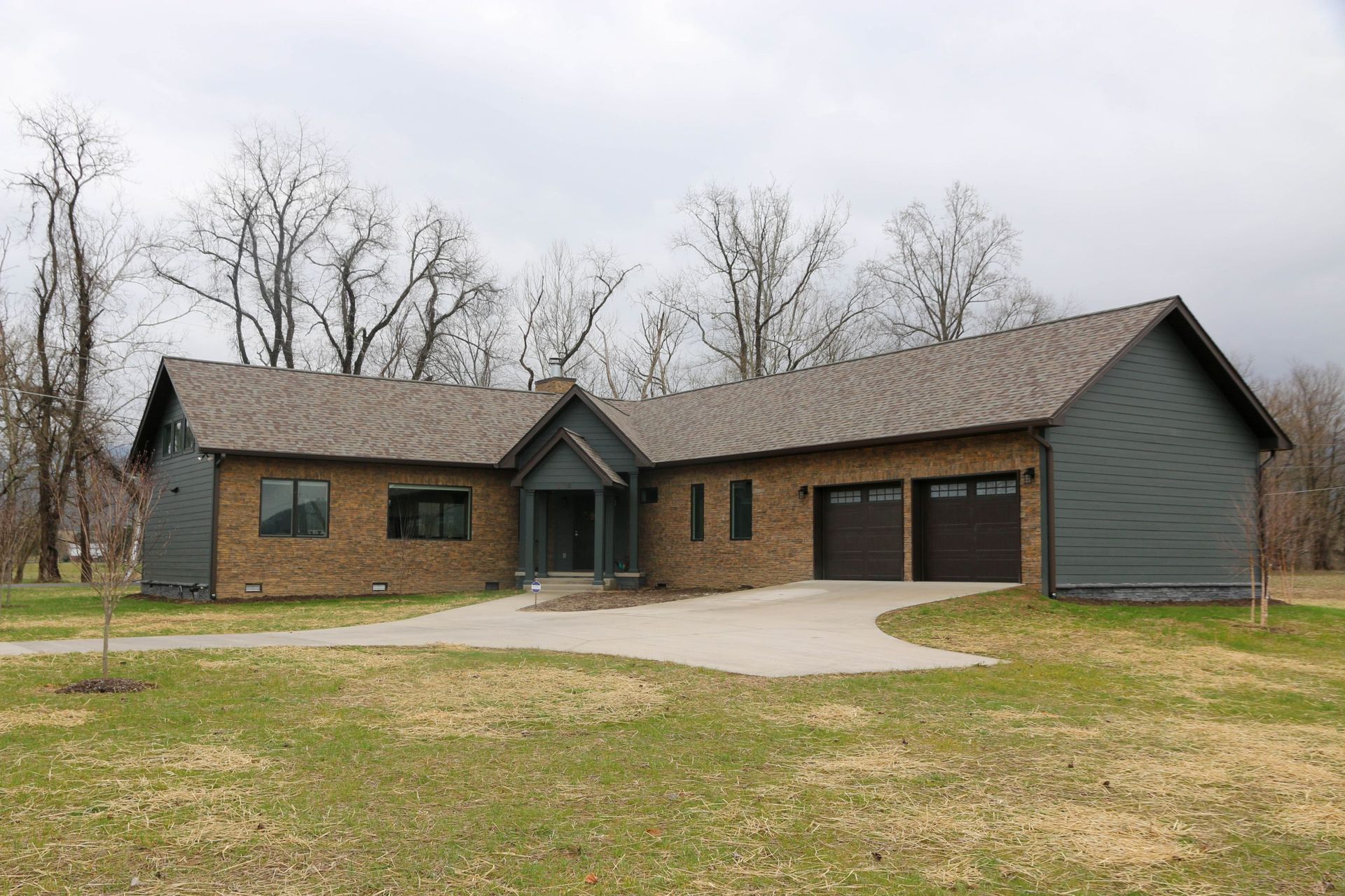A one-story suburban home with brick siding, dark grey accents, a two-car garage, and a driveway in a rural setting.