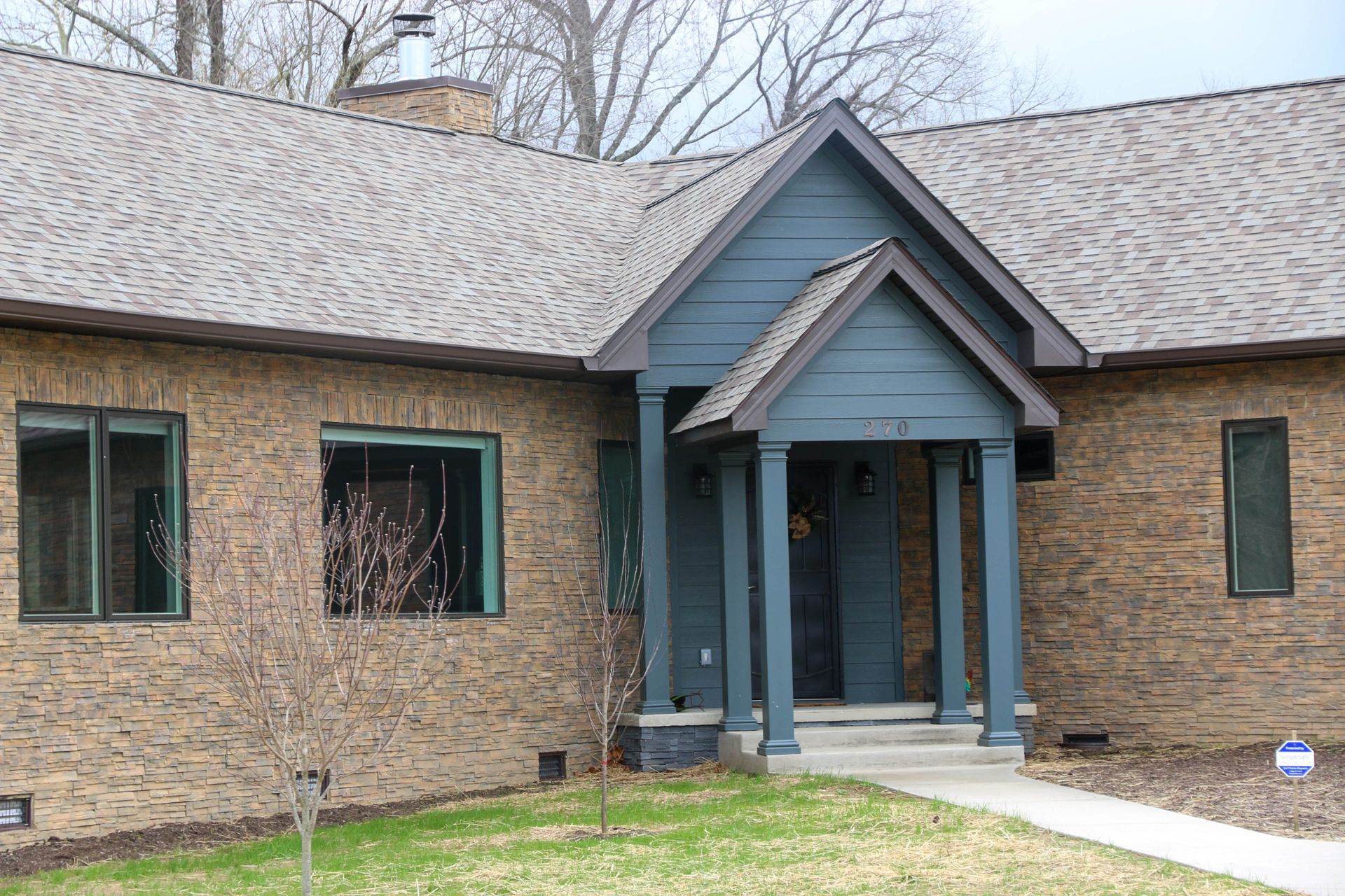 A one-story brick house with a blue front porch entry, dark window frames, and a shingled roof on a grassy lawn.