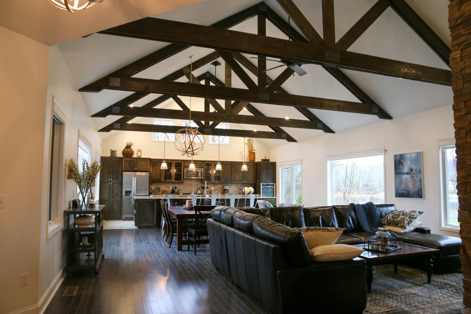 An open-plan living room and kitchen with dark wood ceiling trusses, a leather sectional sofa, and dark hardwood floors.