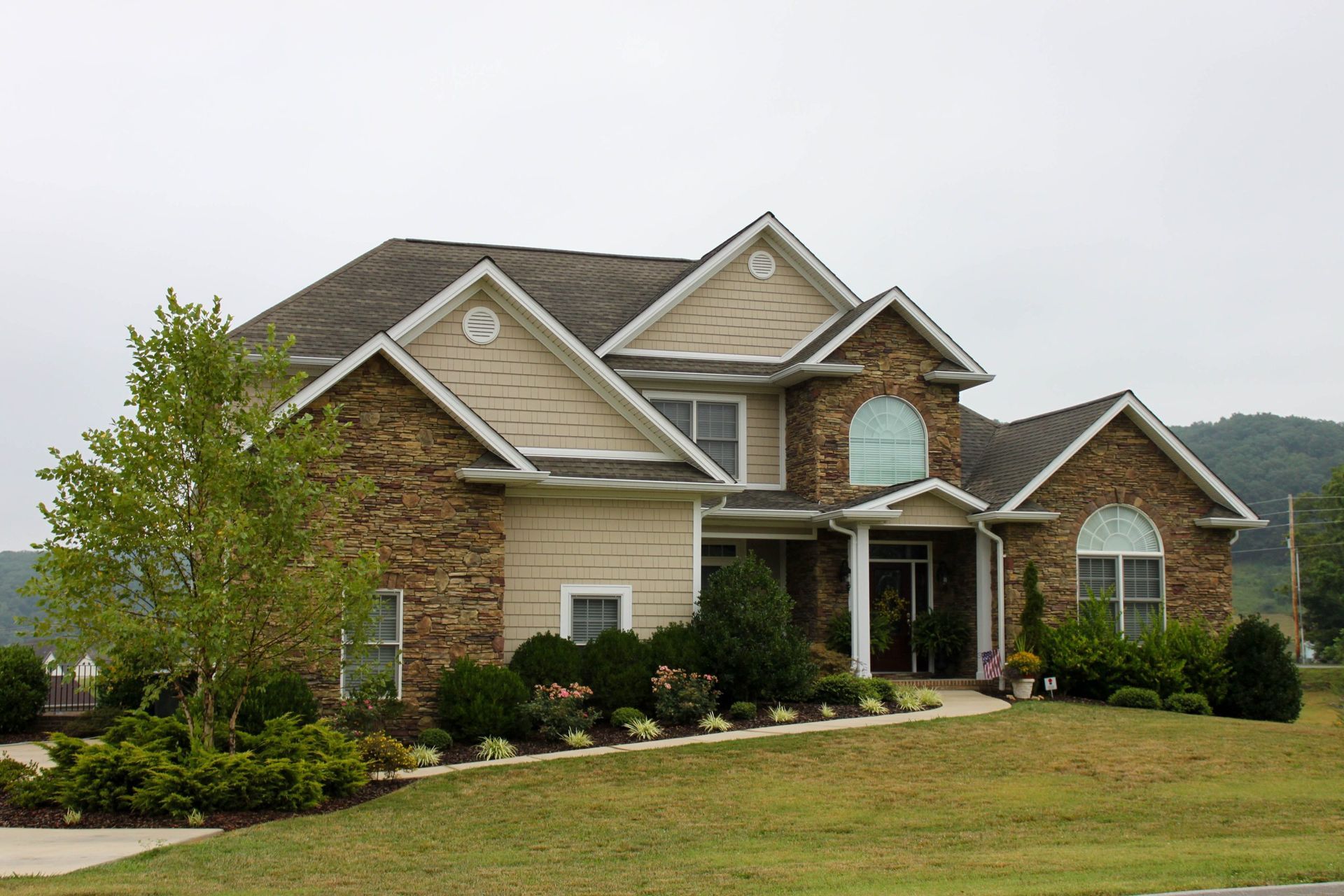 A two-story suburban house with stone and tan siding, gabled rooflines, and a landscaped yard under a cloudy sky.