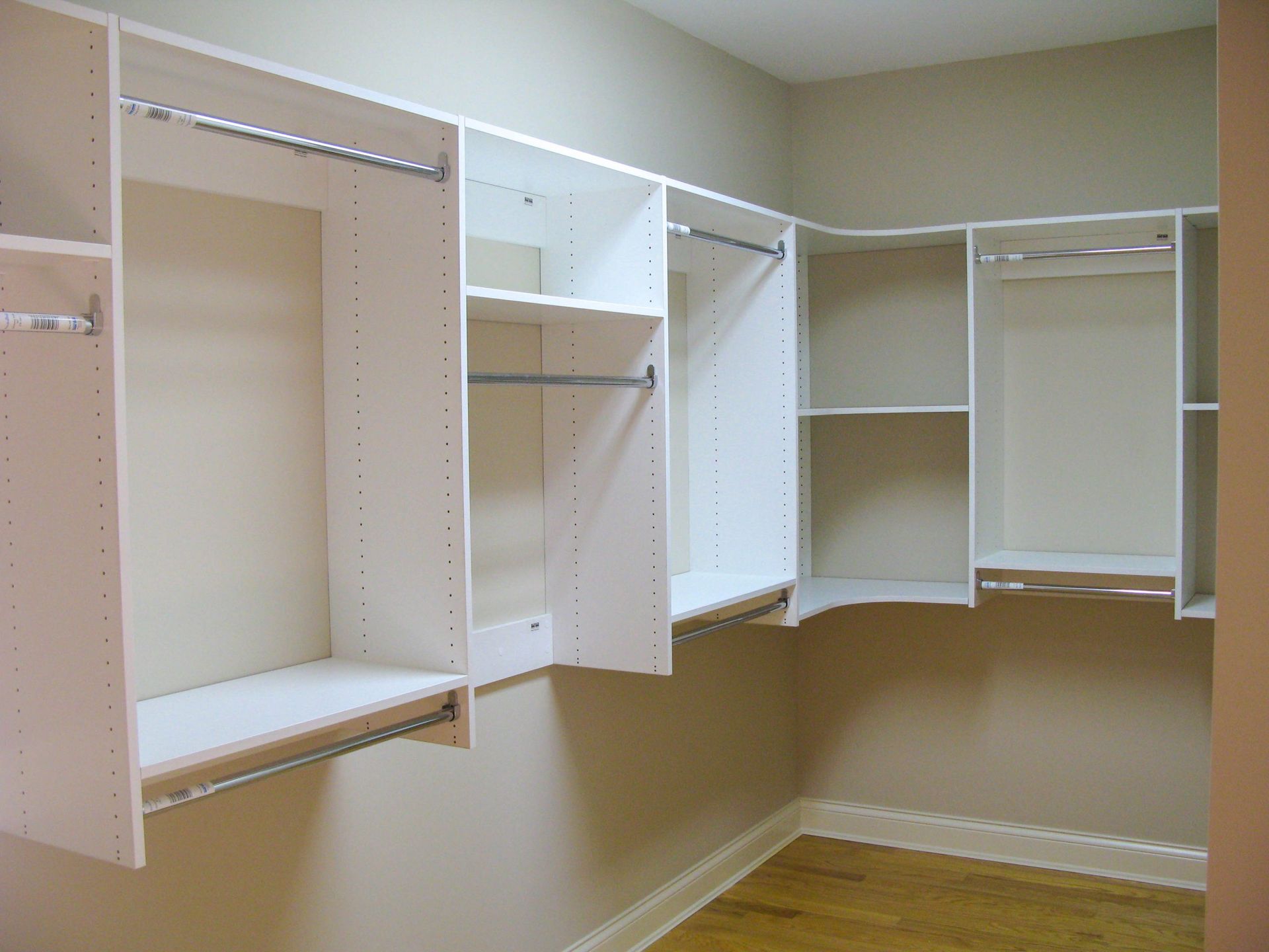 Empty white modular closet shelving units with rods installed along an L-shaped wall over hardwood floors.