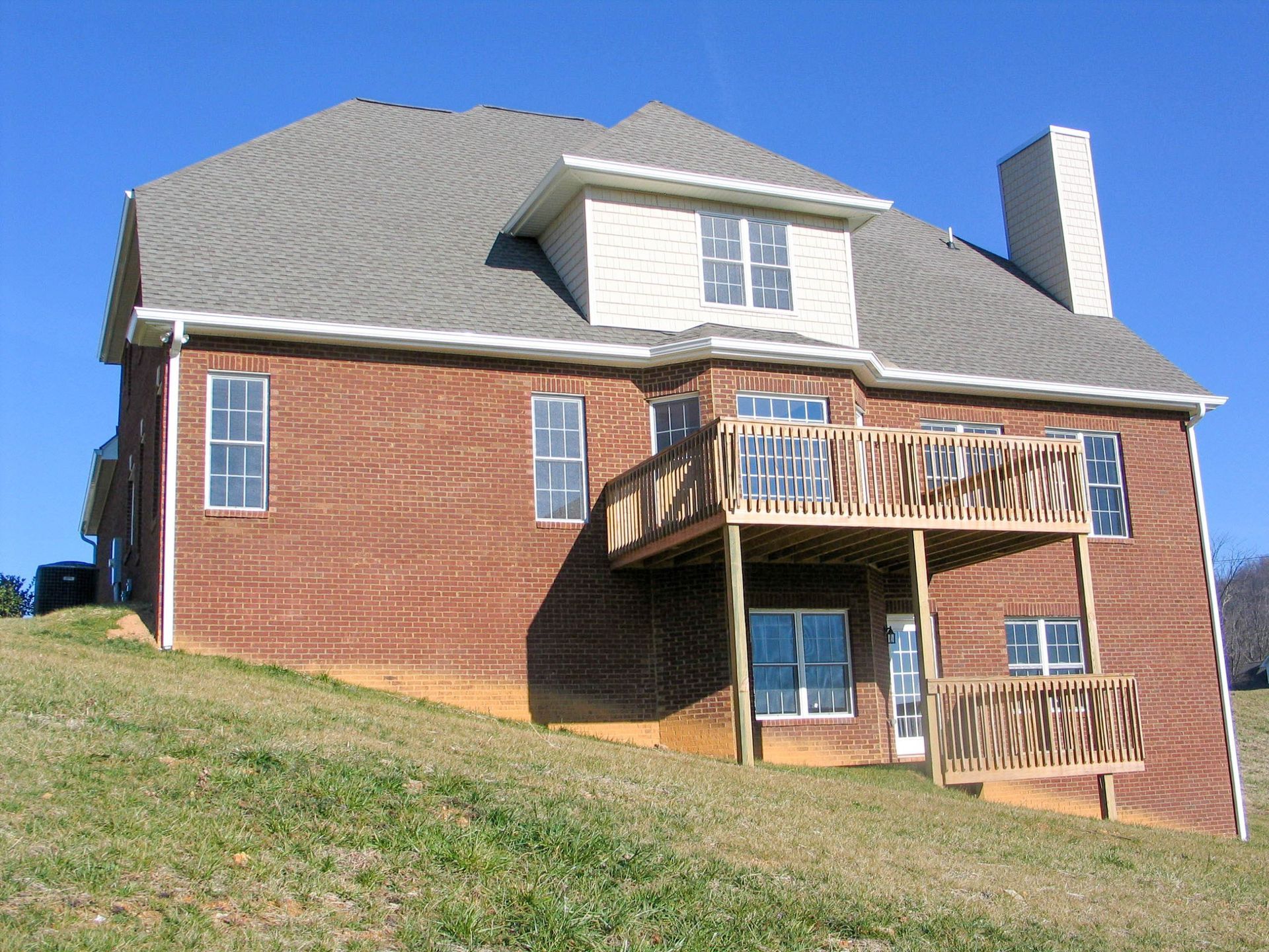 A two-story brick house with a wooden deck and a sloped grassy yard under a clear blue sky.