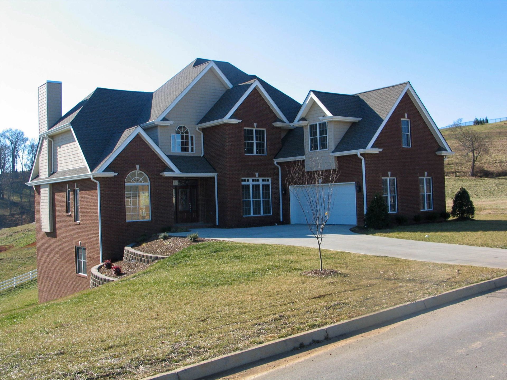 A two-story brick and tan suburban home with an attached garage, situated on a grassy, sloped lot under a clear blue sky.