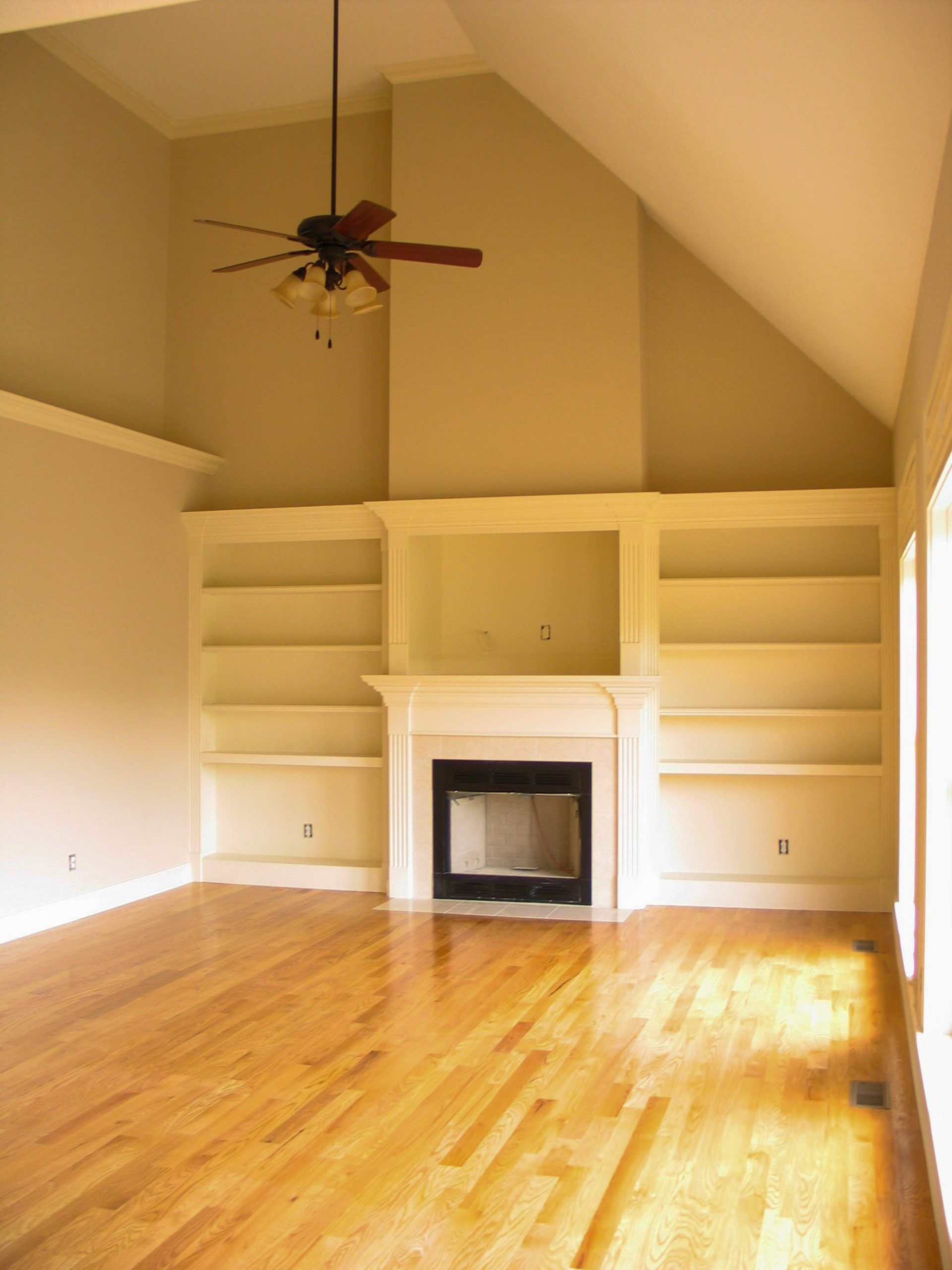 A bright living room with hardwood floors, a vaulted ceiling, a ceiling fan, a fireplace, and built-in bookshelves.