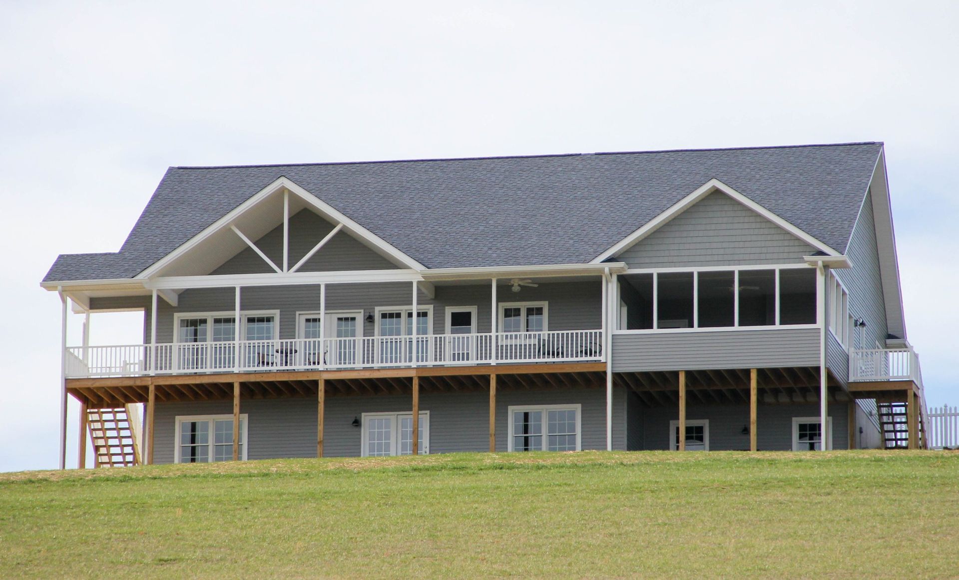 A two-story grey house with a large deck and covered porch sits on a grassy hill under a bright, overcast sky.